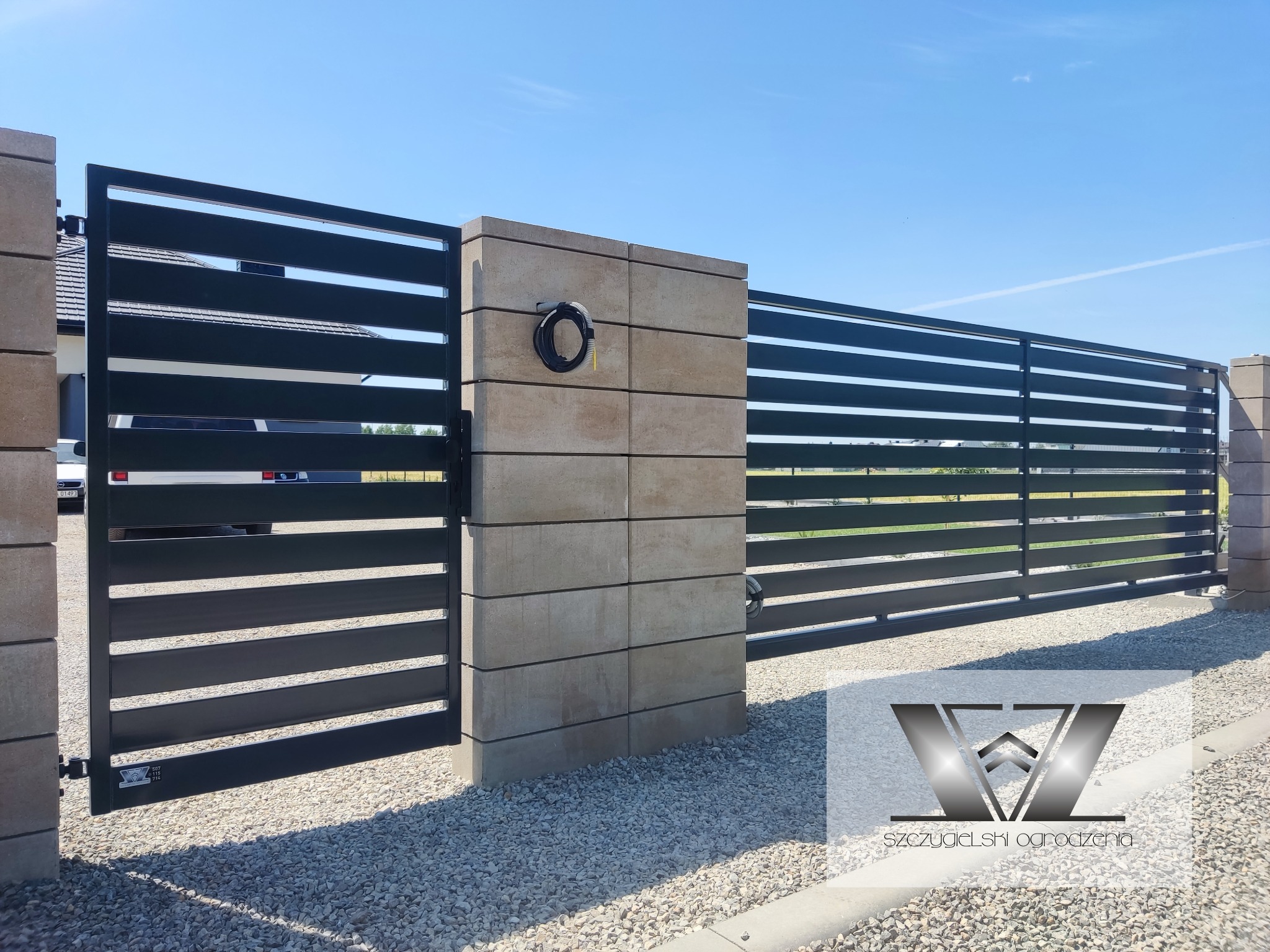 Nowoczesna brama wjazdowa z poziomych, czarnych profili, osadzona na słupkach z szarej kostki, z widocznym przewodem elektrycznym na jednym ze słupków.