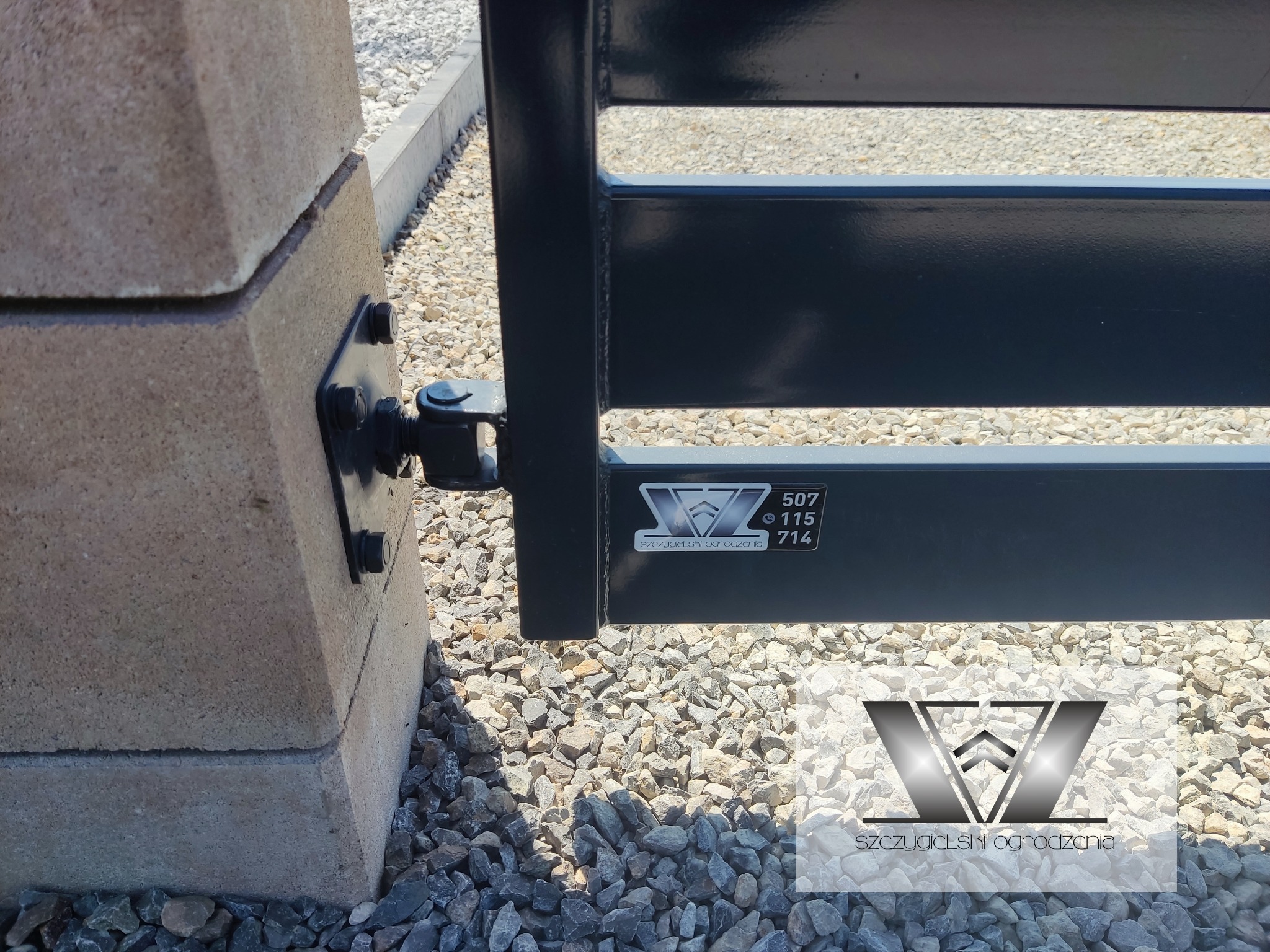 Fragment bramy wjazdowej z metalowymi przęsłami, zamontowanej na betonowym słupku z widocznym zawiasem i logotypem producenta na tle żwirowego podłoża.