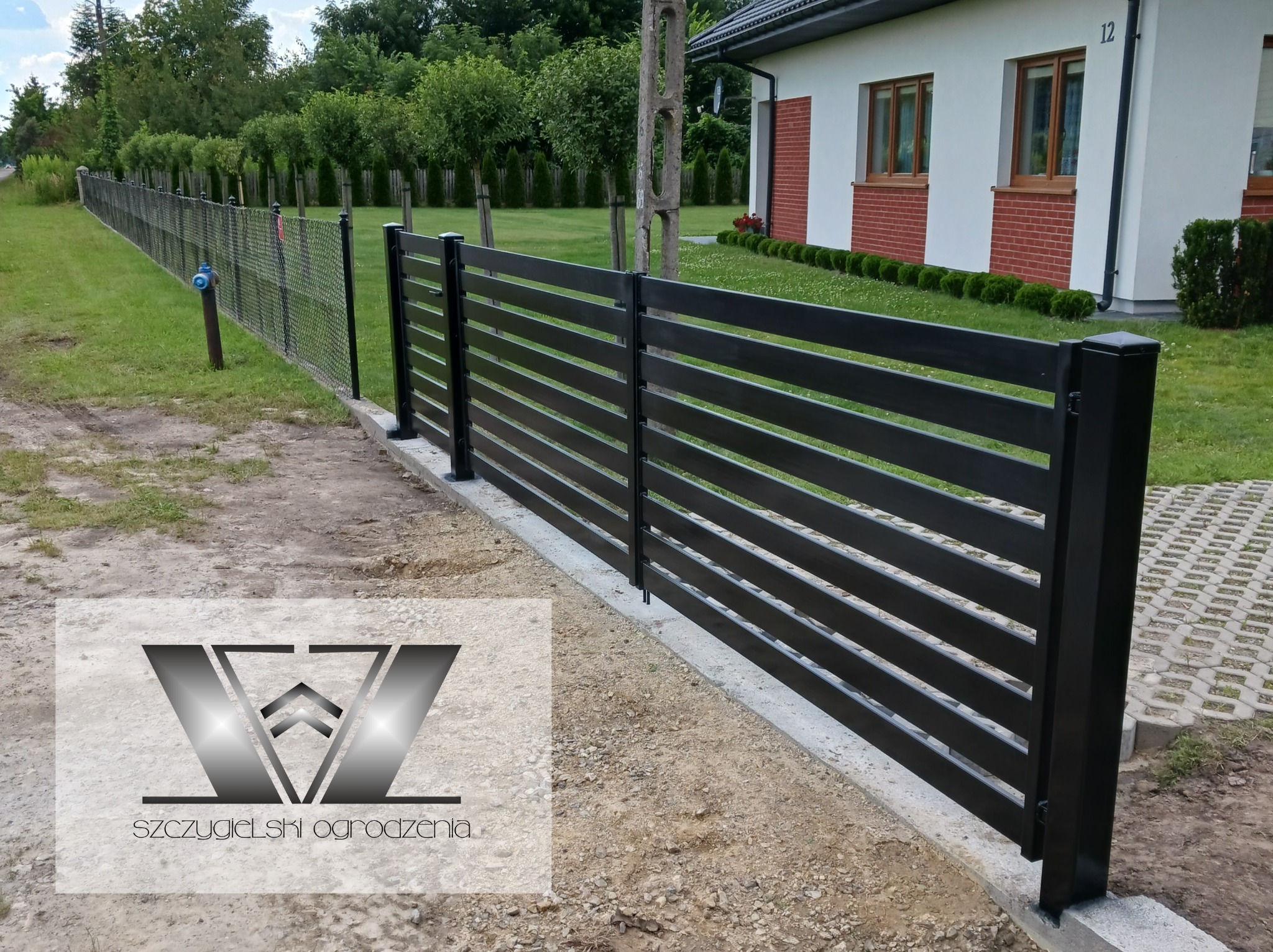Długie, czarne, poziome elementy tworzą nowoczesne ogrodzenie posesji, widoczne logo firmy na pierwszym planie, w tle dom z ceglaną elewacją i zielony trawnik.
