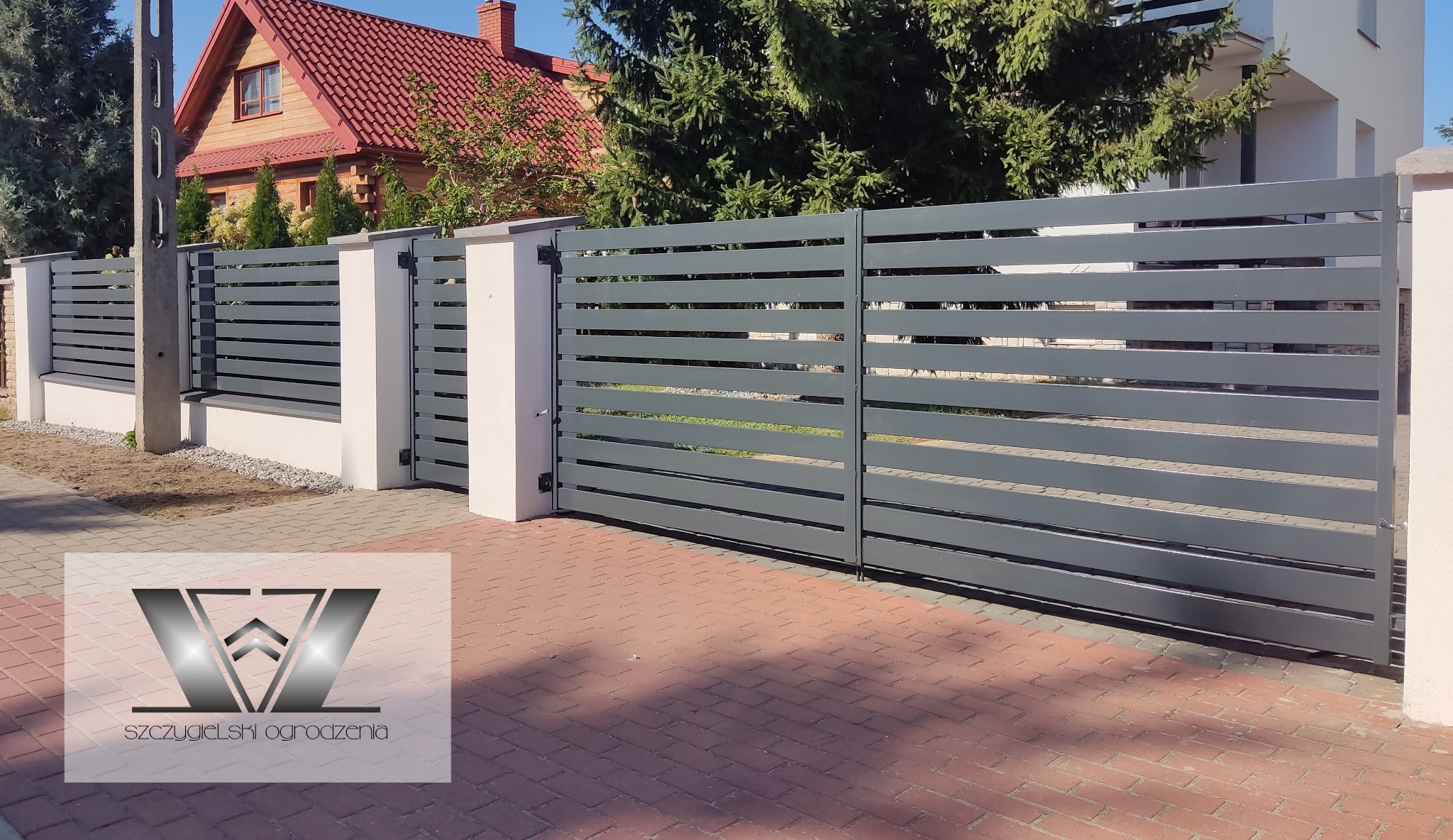 Nowoczesne, szare ogrodzenie panelowe z poziomymi przęsłami i bramą wjazdową, osadzone na białych słupkach, na tle domu z czerwoną dachówką i zielenią.