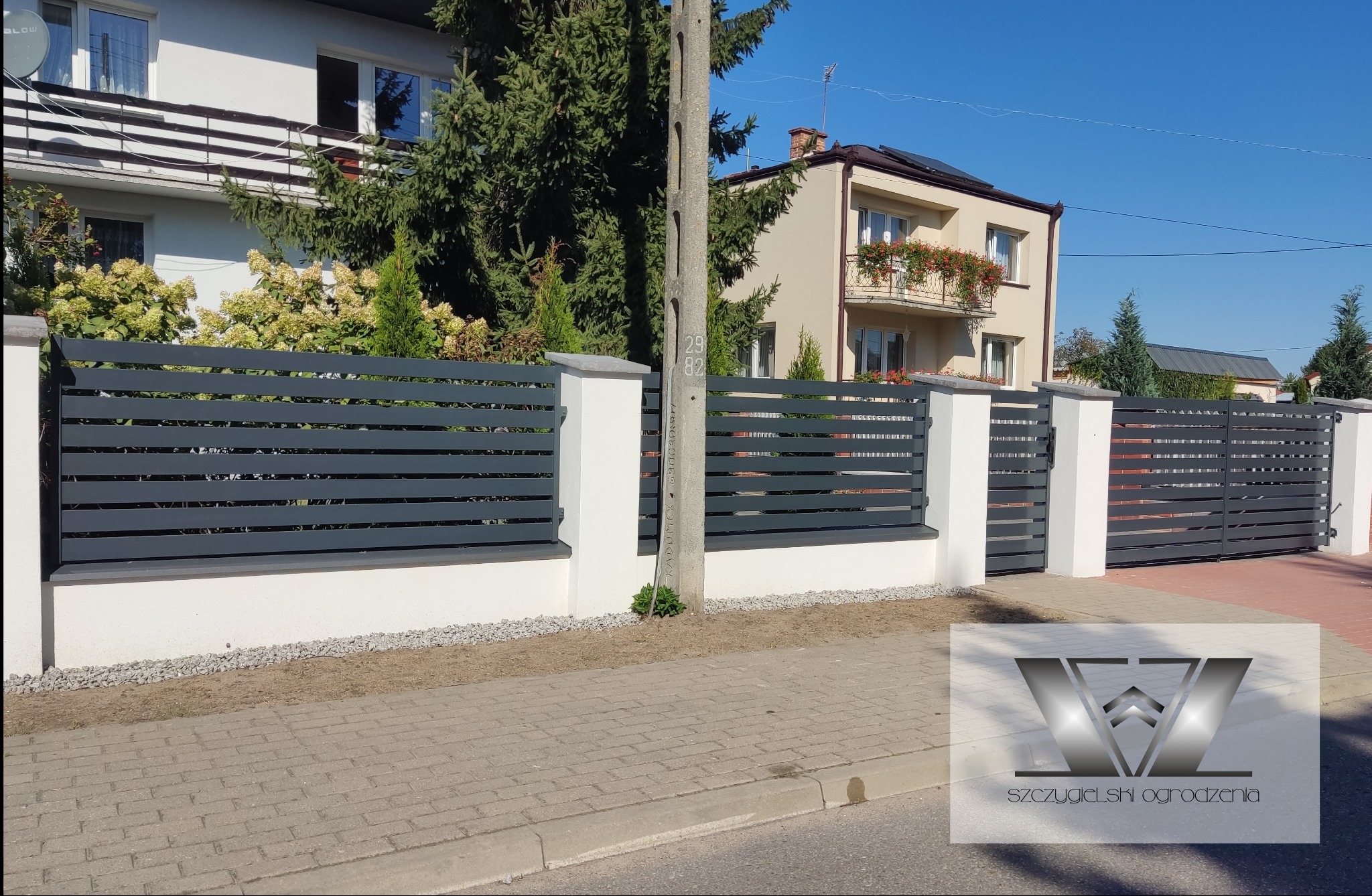 Nowoczesne ogrodzenie z poziomych, szarych paneli zamontowane na białych słupkach, z bramą wjazdową w tle, na tle dwóch domów i słupa energetycznego.