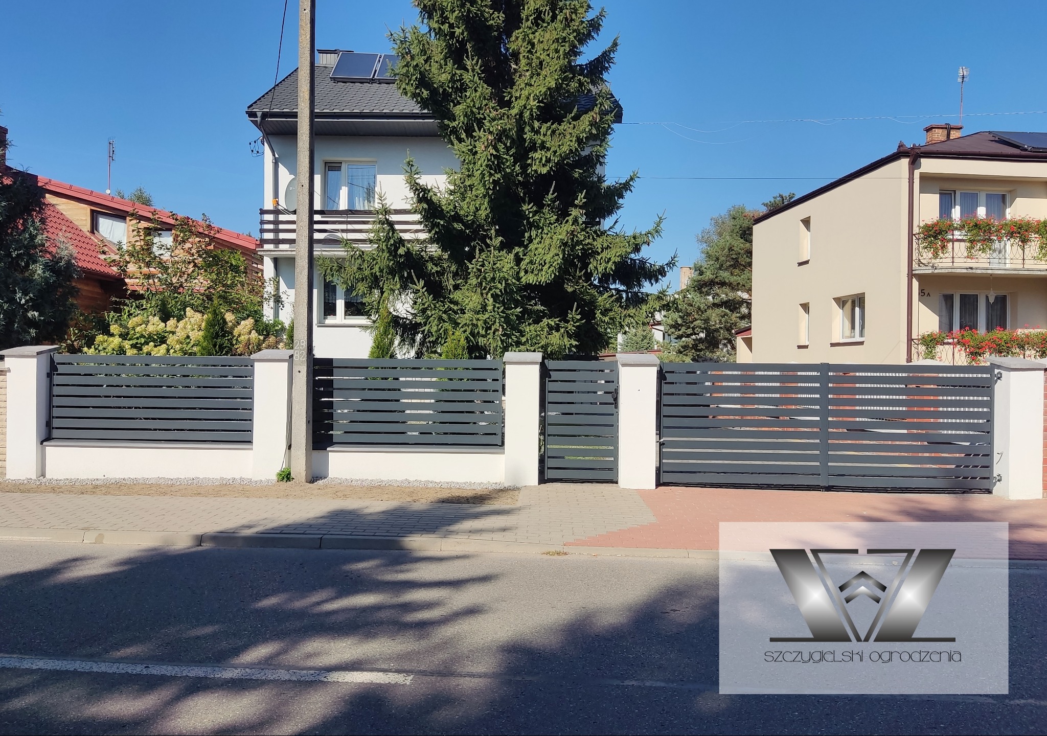Nowoczesne ogrodzenie posesji z poziomych, szarych paneli i białych słupków, brama wjazdowa przesuwna, na tle domów jednorodzinnych w słoneczny dzień.