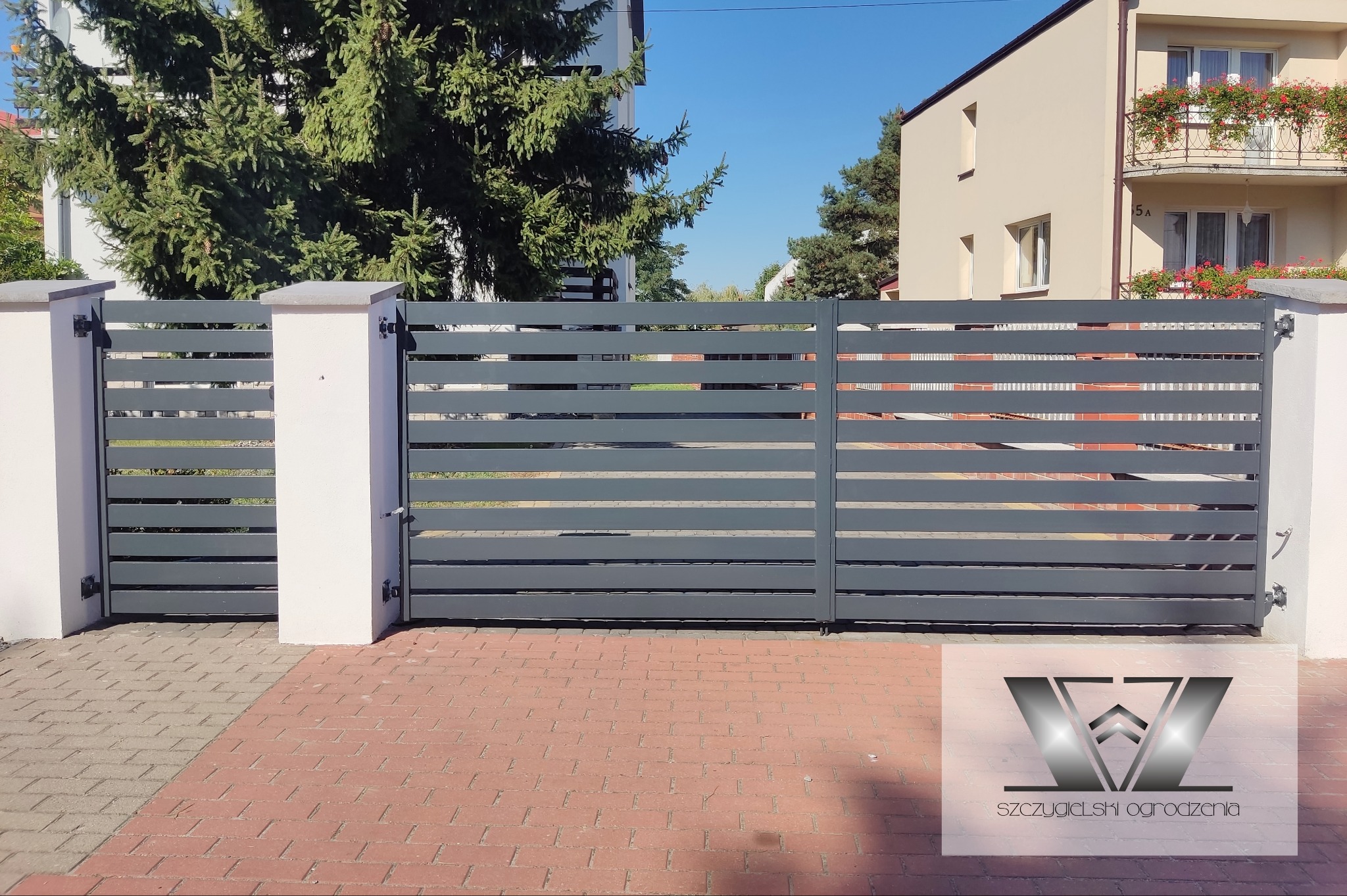 Nowoczesna, pozioma brama panelowa w kolorze grafitowym, zamontowana na białych, kwadratowych słupkach, z widocznym logo firmy w dolnym prawym rogu na tle czerwonej kostki brukowej.
