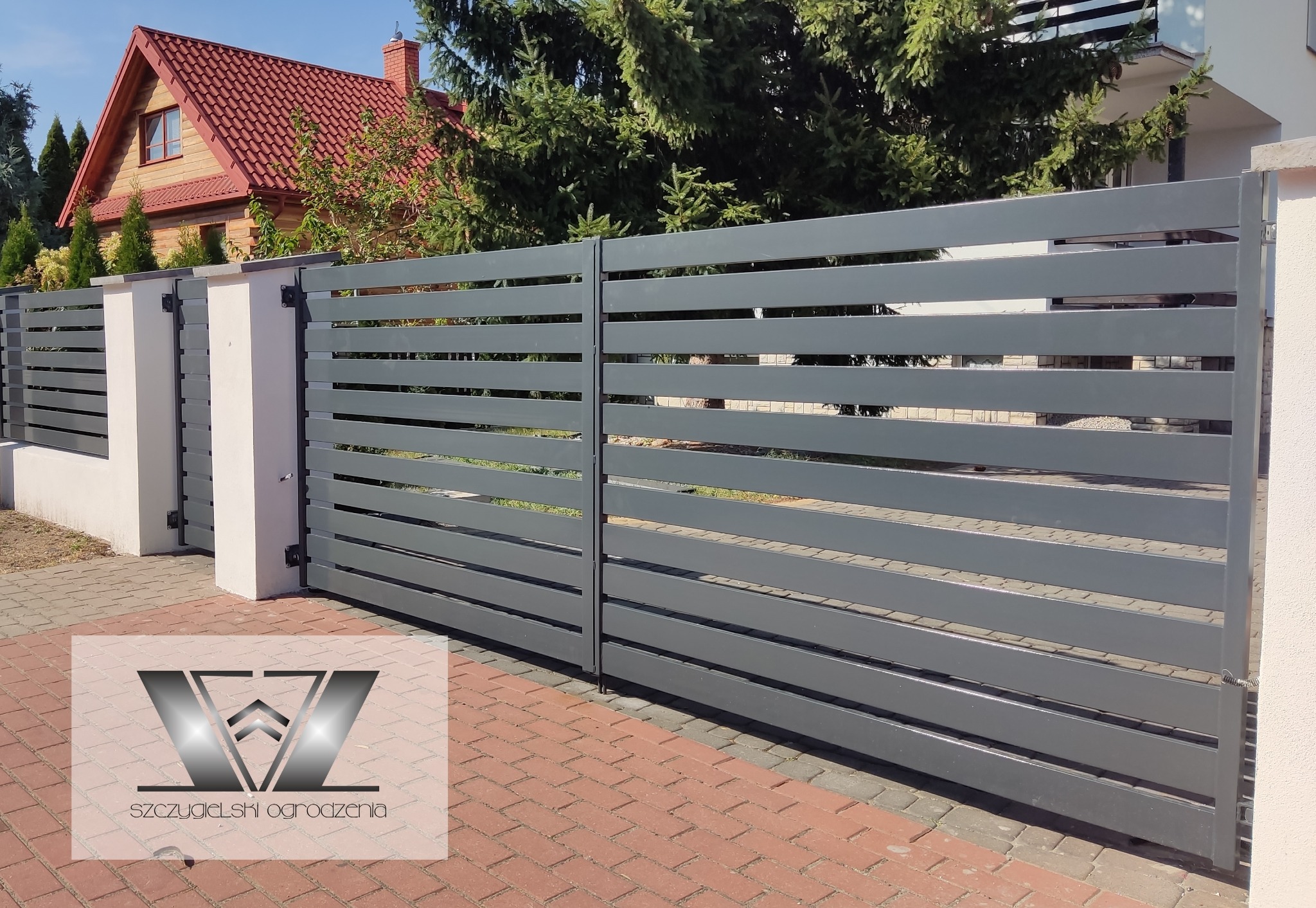 Nowoczesna brama wjazdowa z poziomych, szarych paneli, zamontowana na białych słupkach, z widocznym fragmentem domu z czerwoną dachówką w tle.