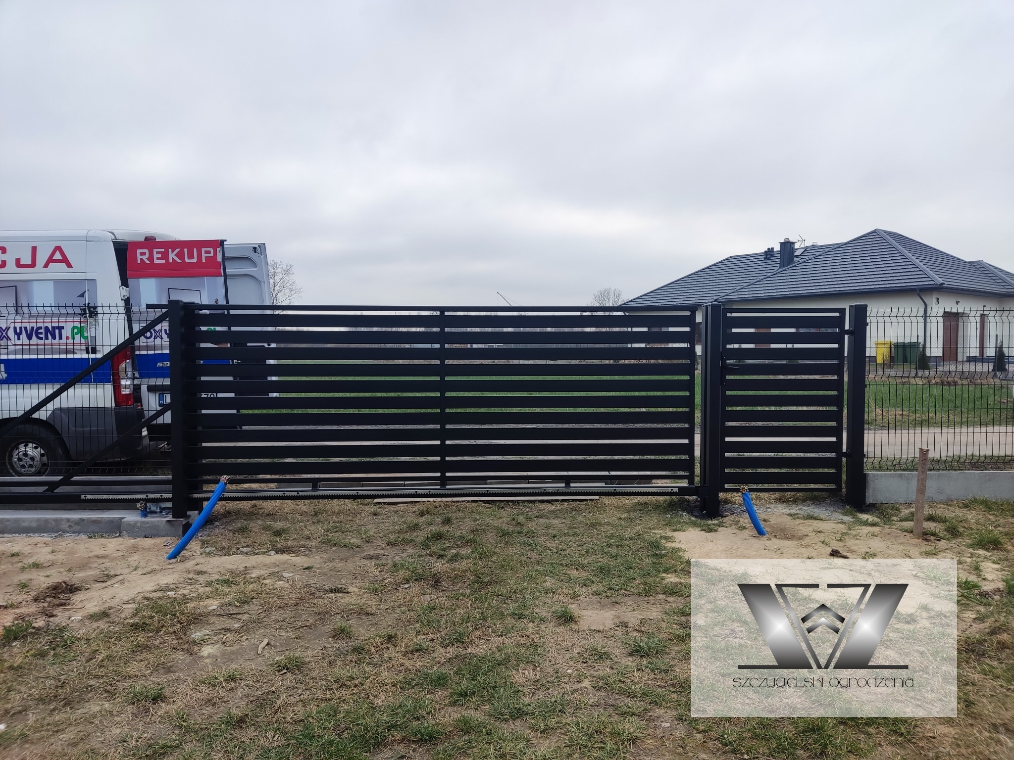 Nowoczesna, pozioma brama przesuwna w kolorze czarnym, z widocznym mechanizmem automatyki i niebieską rurą ochronną kabli, na tle szarego nieba i częściowo widocznego samochodu dostawczego...