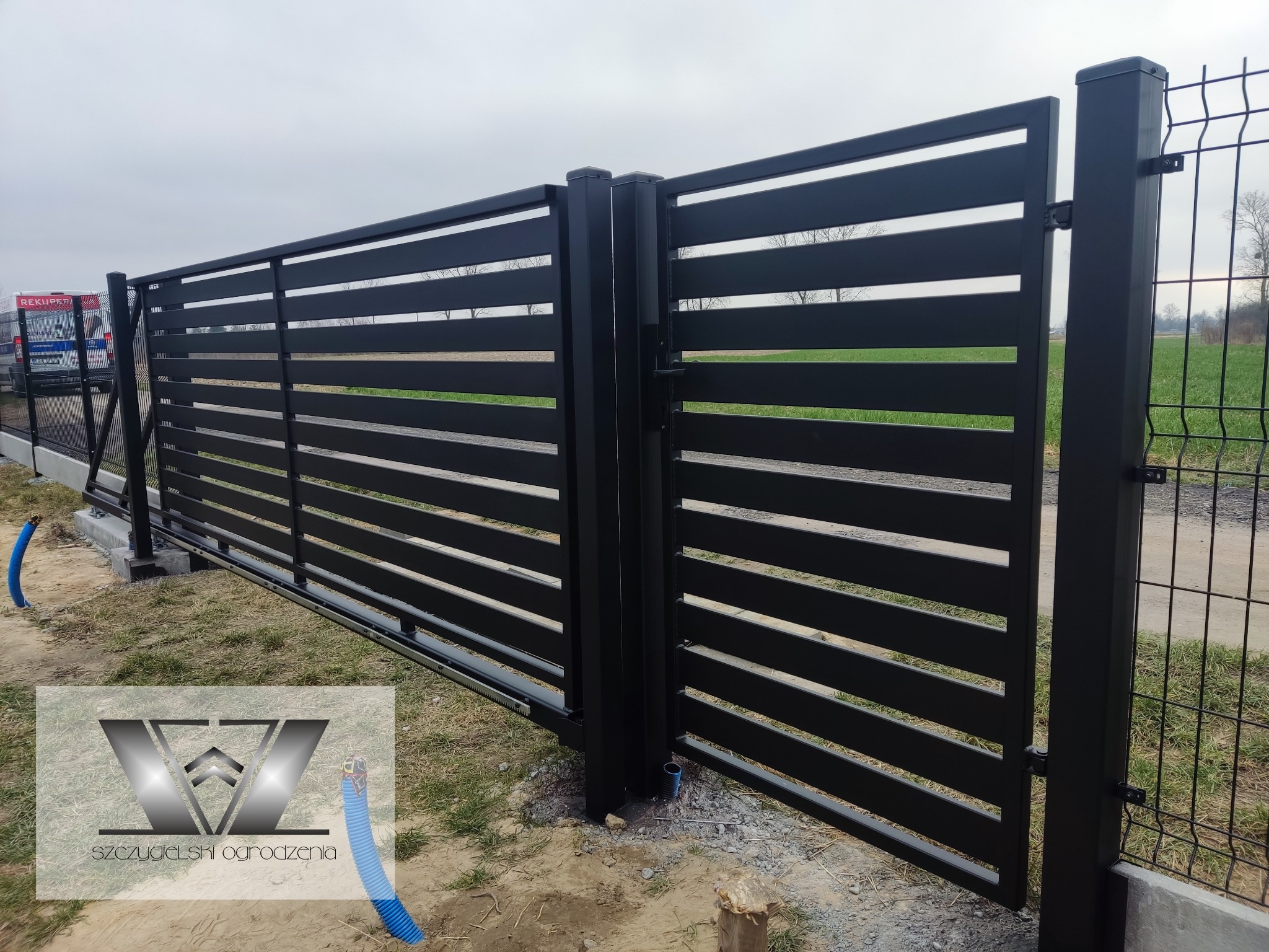 Nowoczesna, przesuwna brama wjazdowa z poziomych, czarnych profili, widoczne elementy automatyki i peszel z kablem, w tle ogrodzenie panelowe.