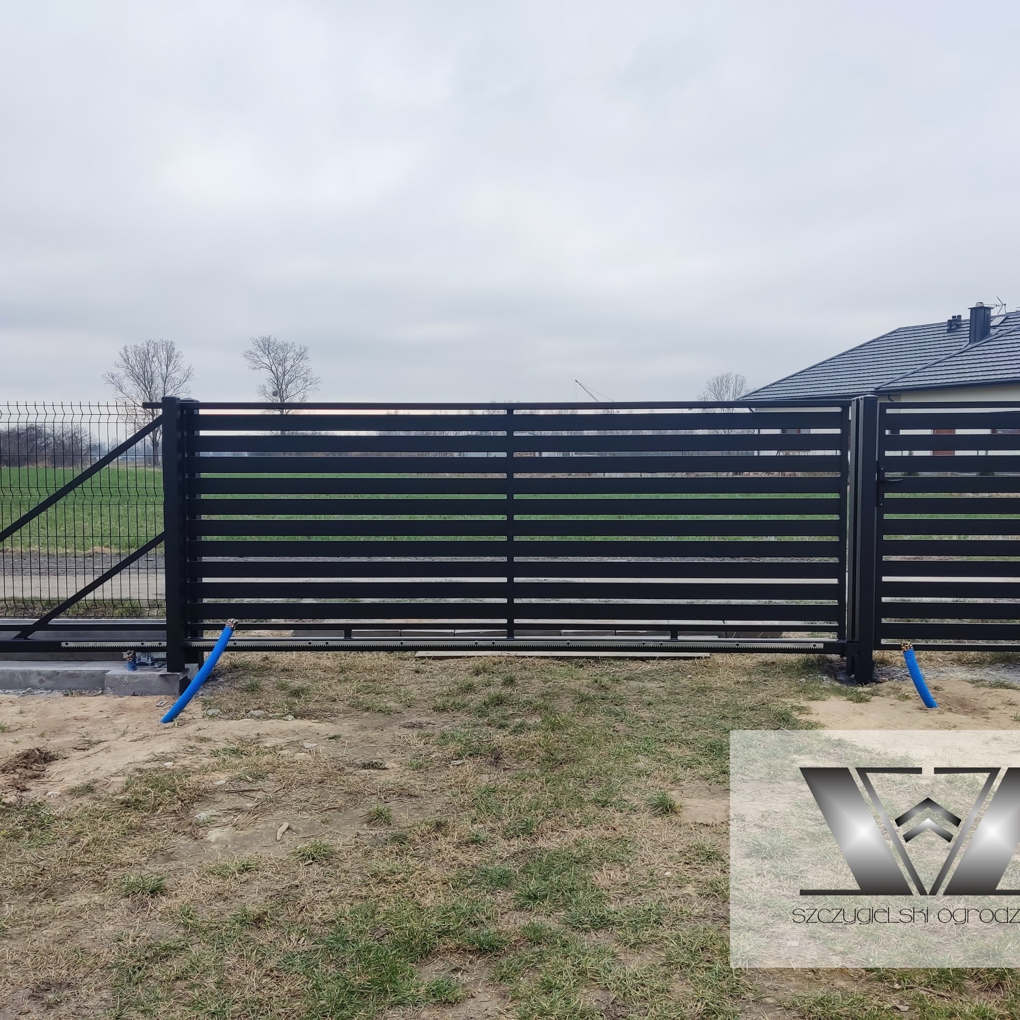 Nowoczesna brama przesuwna z poziomych profili, czarna, z widocznym mechanizmem, niebieski peszel doprowadzony do napędu, w tle ogrodzenie z siatki i dom.