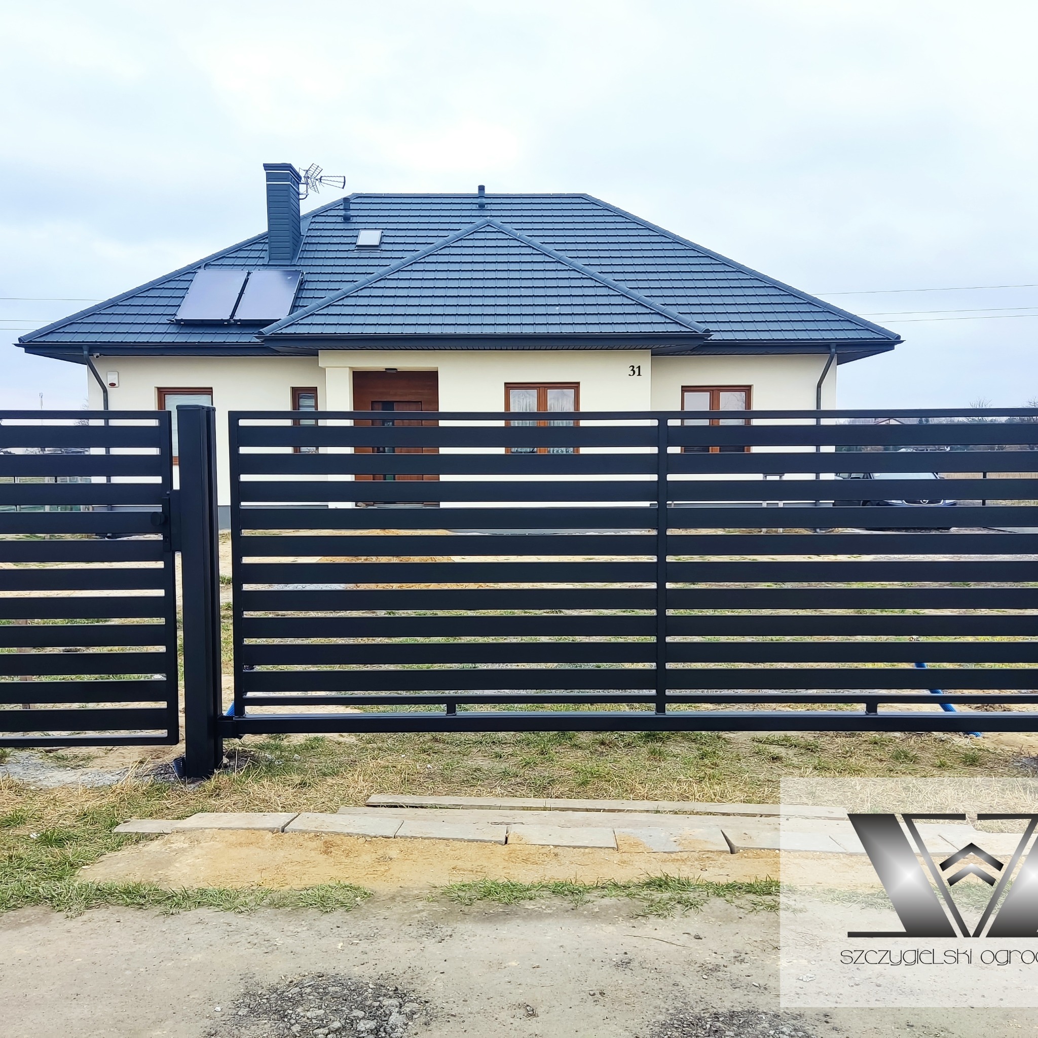 Nowoczesne, poziome, czarne przęsła ogrodzeniowe na tle domu z szarą dachówką i panelami słonecznymi.