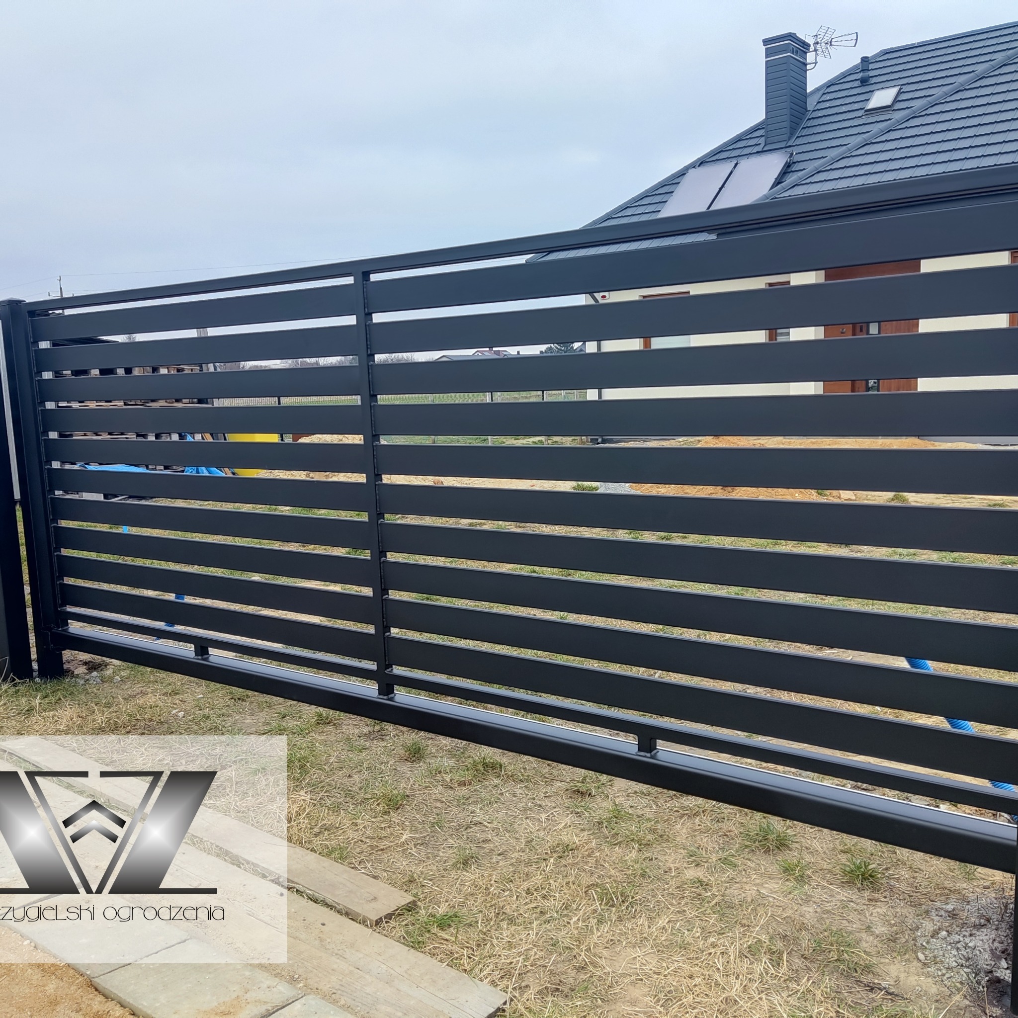 Nowoczesne, poziome ogrodzenie panelowe w kolorze antracytowym, z widocznym domem w tle i elementami logo firmy w lewym dolnym rogu.