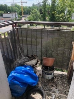 Balkon przed sprzątaniem: widoczne gruz, worki z odpadami, doniczka i wiadro, zarośnięta balustrada, w tle drzewa i żuraw budowlany.