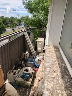 Zabrudzony balkon przed sprzątaniem, widoczne resztki budowlane, gruz, stare farby i narzędzia, zarośnięte otoczenie za barierką.