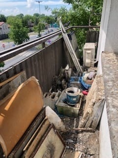 Zabrudzony balkon z gruzem, starymi ramami obrazów, wiadrem po farbie, pustakami i innymi odpadami budowlanymi. W tle widoczna ulica z samochodami i drzewa.