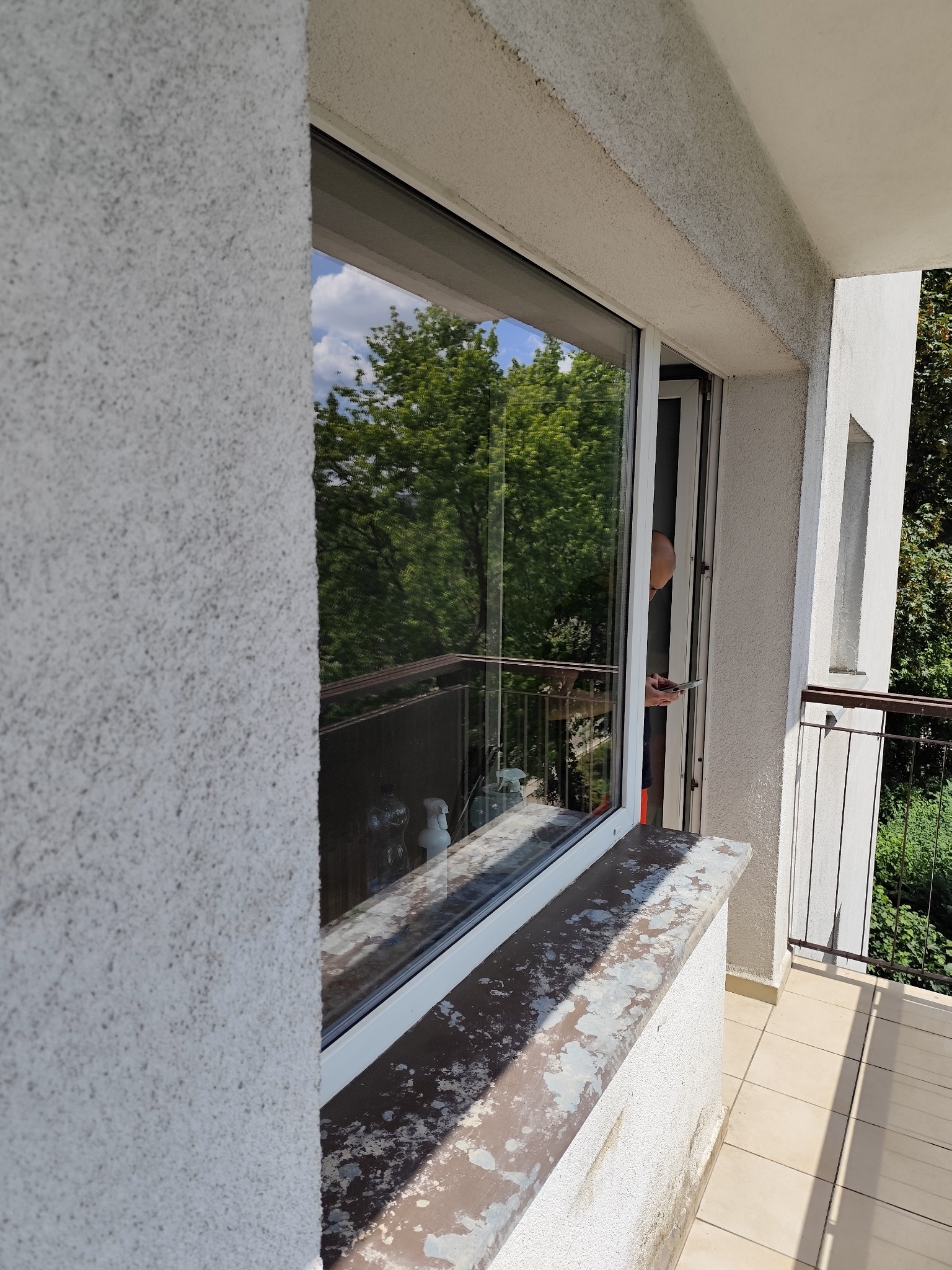 Okno balkonowe z widokiem na zieleń, brudny parapet zewnętrzny, osoba w drzwiach balkonowych z telefonem.