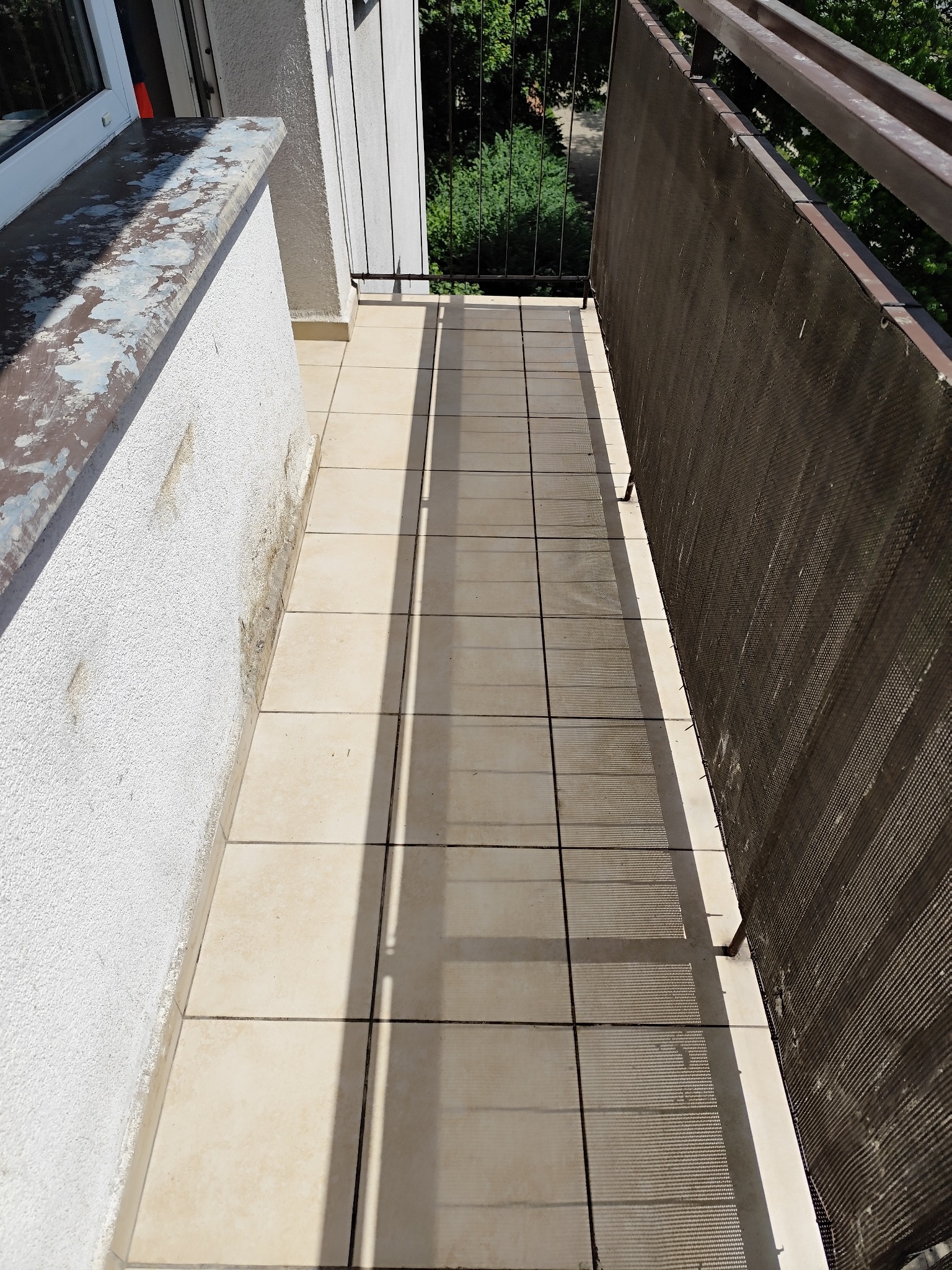 Balkon w Łodzi z widocznymi śladami zabrudzeń na ścianie i płytkach podłogowych, osłonięty brązową siatką. Widoczne także odpryski farby na parapecie.
