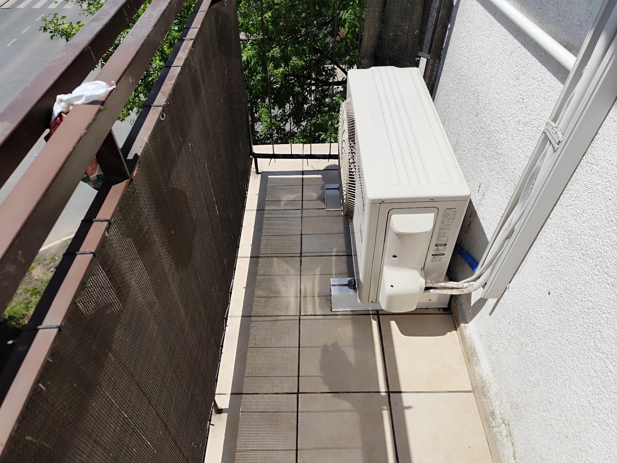 Biały klimatyzator zamontowany na balkonie z brązową balustradą i perforowaną osłoną, widok z góry na ulicę.