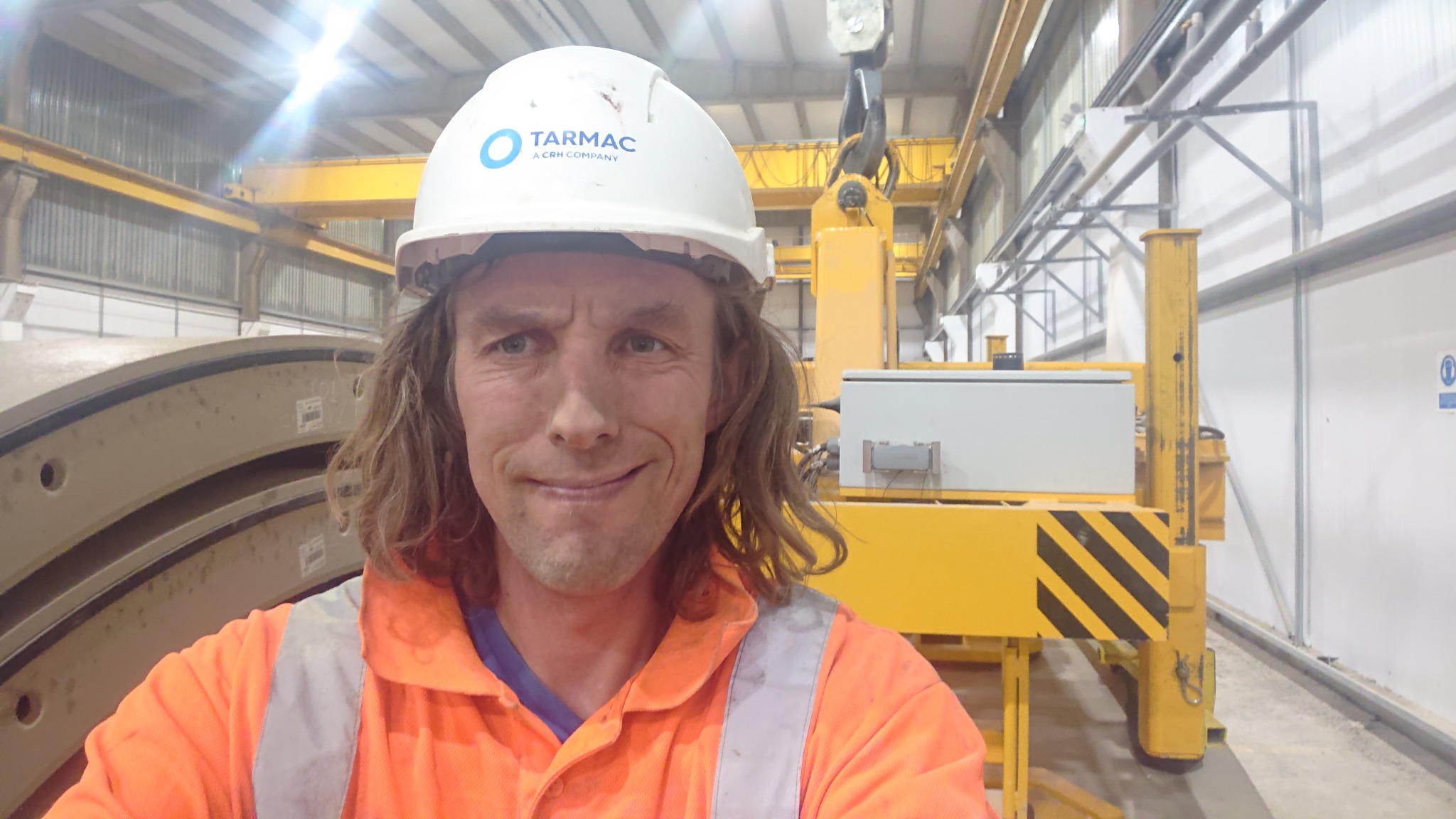 Pracownik w kasku Tarmac i pomarańczowej kamizelce odblaskowej, na tle suwnicy i betonowych elementów konstrukcyjnych w hali przemysłowej.