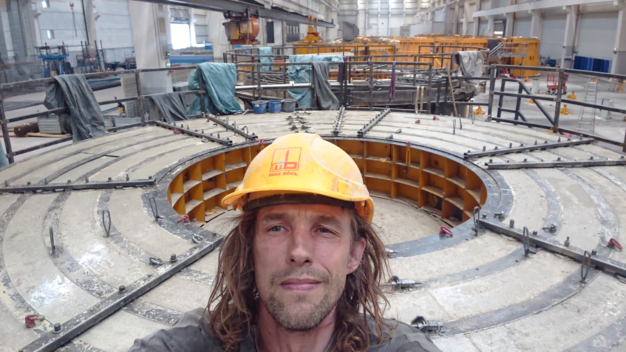 Pracownik w kasku Max Bögl na tle okrągłej konstrukcji szalunkowej w hali przemysłowej, widoczne elementy stalowe i betonowe.