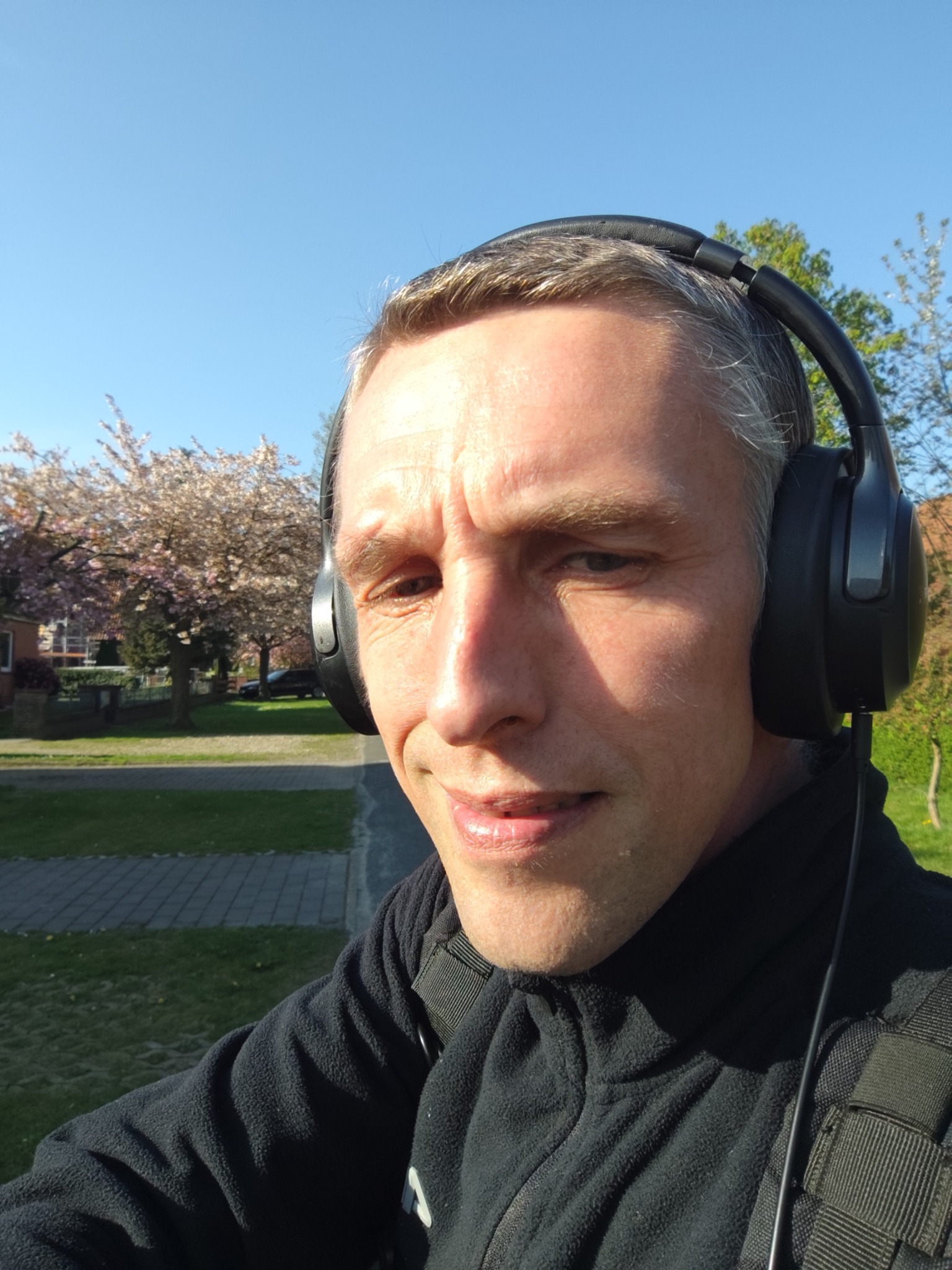 Mężczyzna w słuchawkach na tle kwitnących drzew wiśni. Słoneczny dzień, widok na park i budynki w tle. Selfie z bliska, naturalne światło.