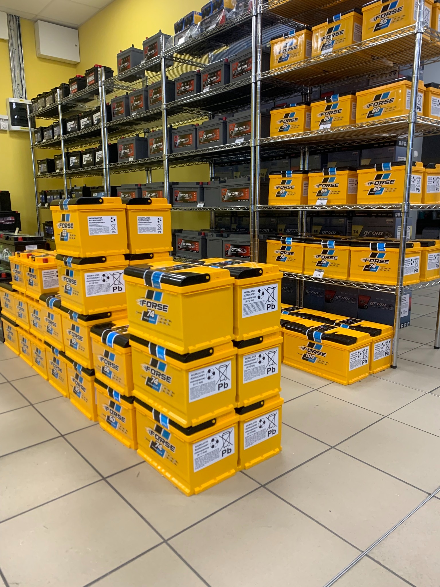 Składowisko akumulatorów samochodowych marki Forse, różnych modeli i pojemności, ułożonych w stosy i na metalowych regałach w magazynie sklepu motoryzacyjnego.