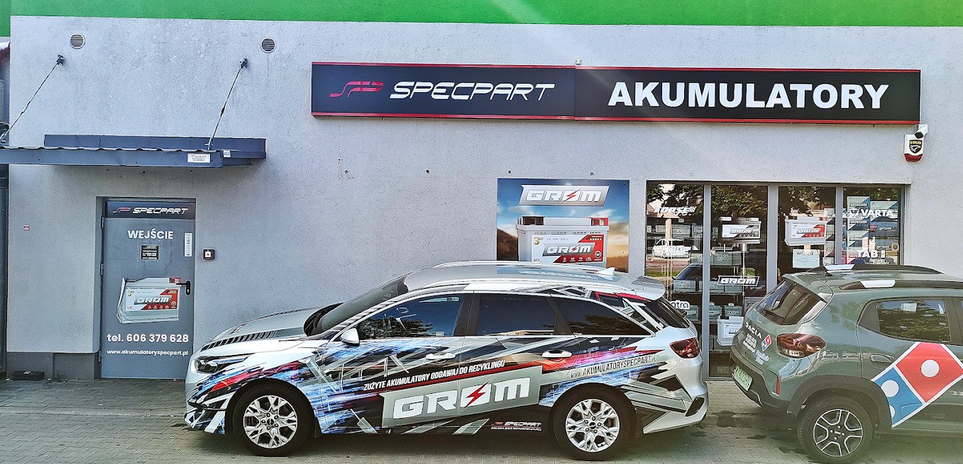 Fasada budynku Specpart Akumulatory z szyldem, wejściem i reklamami akumulatorów Grom, obok zaparkowane dwa samochody, jeden oklejony grafiką reklamową firmy. Widoczny numer telefonu i adres strony...