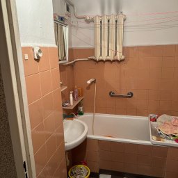 ROJO Radosław Otfinowski - Wnętrze zaniedbanej łazienki z brązowymi płytkami, białą wanną i umywalką, widoczny stary grzejnik, pranie suszące się pod sufitem i wiadro ze śmieciami