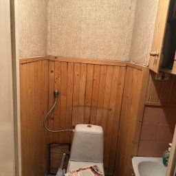 ROJO Radosław Otfinowski - Niefachowo wykończona łazienka z drewnianą boazerią, tapetą imitującą płótno, niedokończonymi fugami, stertą gazet na sedesie i widocznym brakiem porządku.