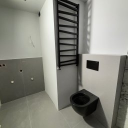 ROJO Radosław Otfinowski - Częściowo wykończona łazienka z czarną wiszącą toaletą, grzejnikiem drabinkowym i szarymi płytkami, widoczne niedokończone instalacje wodne.