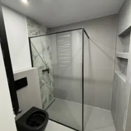 Nowoczesna łazienka w odcieniach szarości z czarną armaturą, kabiną prysznicową typu walk-in z marmurową ścianą i podtynkową toaletą.