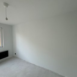 ROJO Radosław Otfinowski - Pusty, jasny pokój z białymi ścianami, betonową podłogą i czarnym grzejnikiem pod oknem z widokiem na drzewa; surowy stan wykończenia przed umeblowaniem.