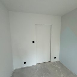 ROJO Radosław Otfinowski - Pomalowane na biało wnętrze pokoju z ukrytymi drzwiami i szarą podłogą, fragment ściany w odcieniach błękitu z geometrycznym wzorem w rogu.