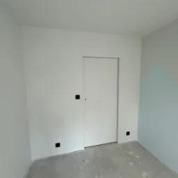 Pomalowane na biało wnętrze pokoju z ukrytymi drzwiami i szarą podłogą, fragment ściany w odcieniach błękitu z geometrycznym wzorem w rogu.