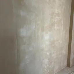 Ściana po nałożeniu pierwszej warstwy tynku w odcieniach beżu, widoczne nierówności i surowy stan wykończenia, z otworem na instalację elektryczną w dolnej części.