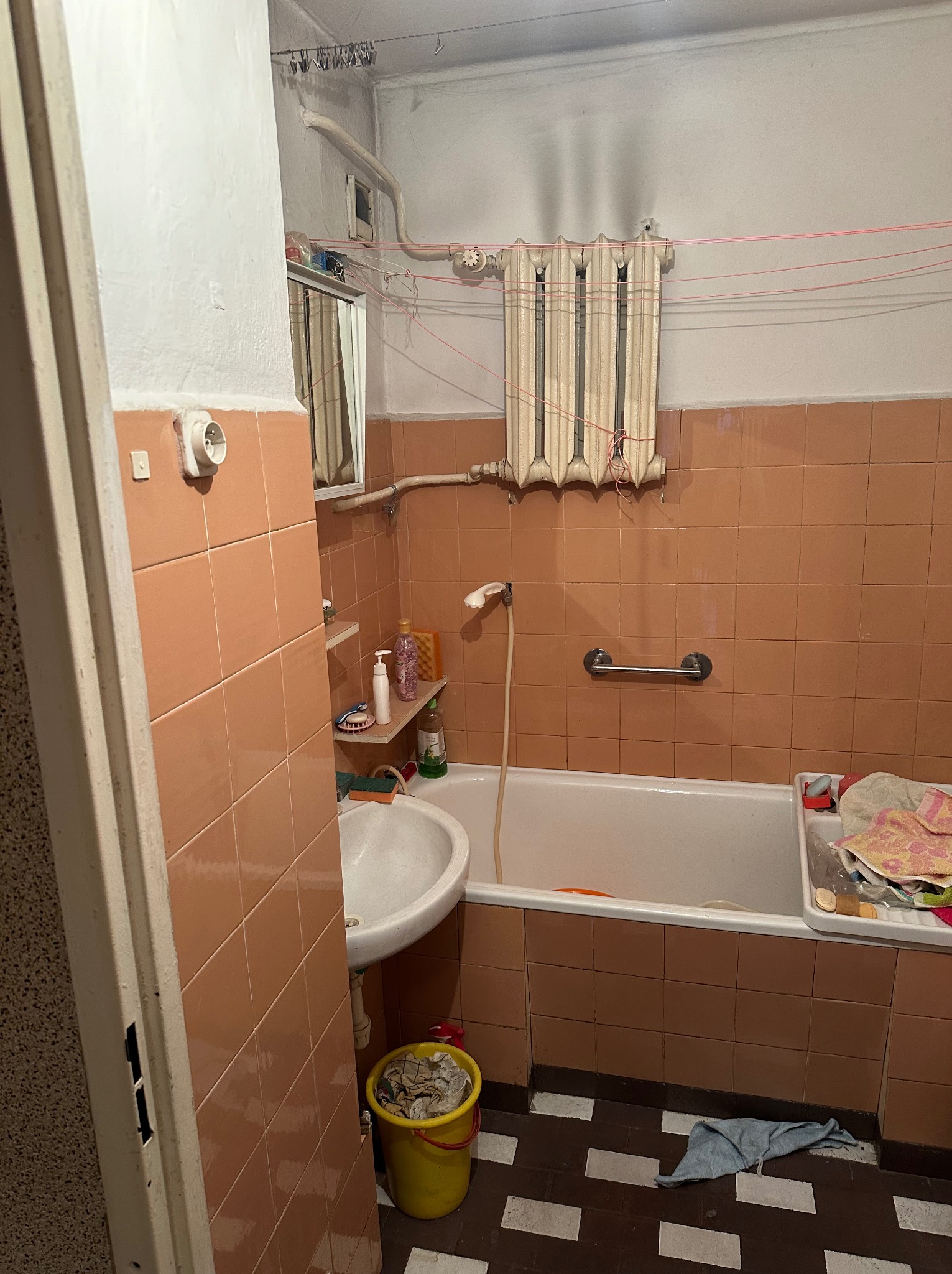 Wnętrze zaniedbanej łazienki z brązowymi płytkami, białą wanną i umywalką, widoczny stary grzejnik, pranie suszące się pod sufitem i wiadro ze śmieciami