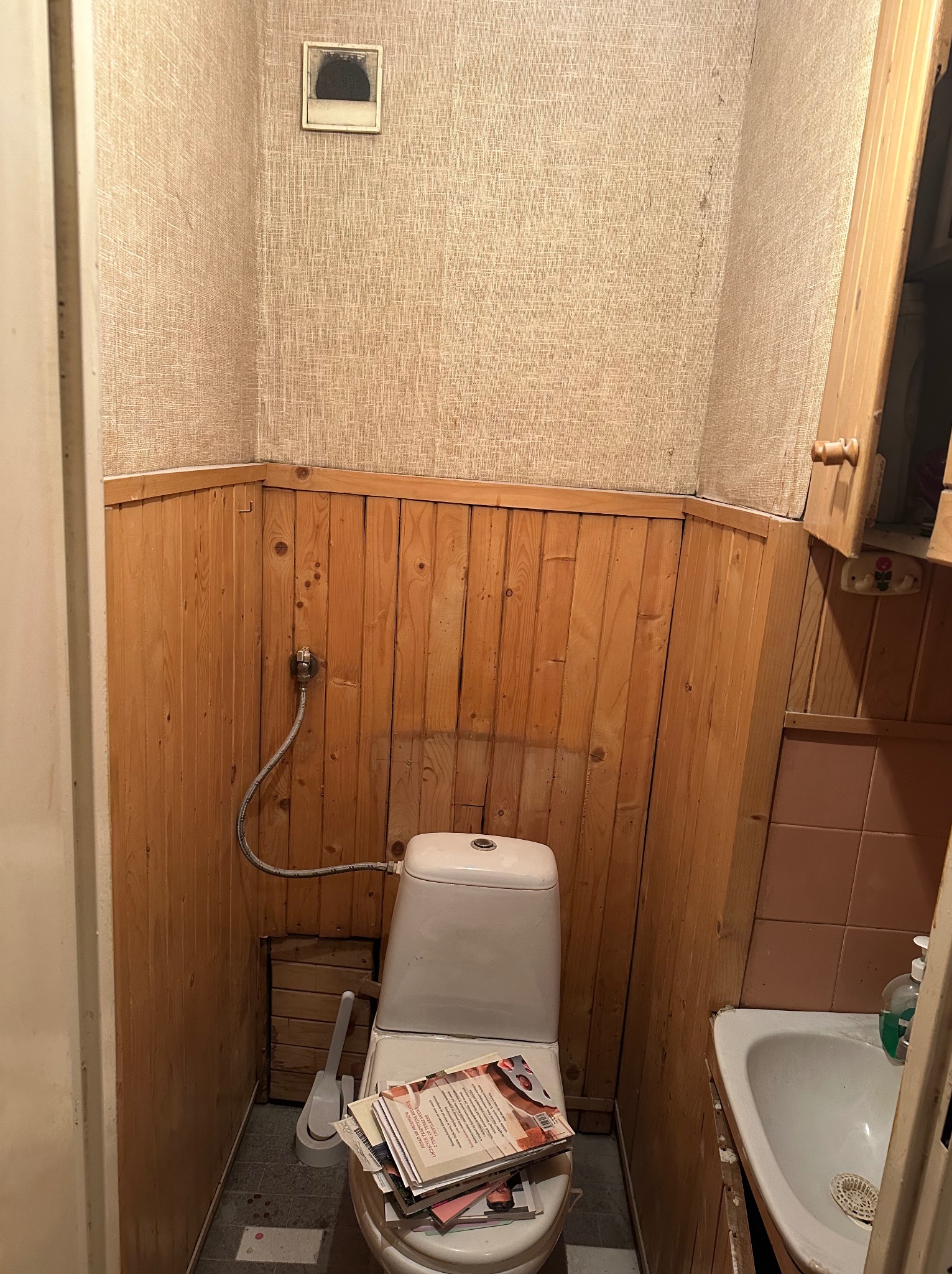 Niefachowo wykończona łazienka z drewnianą boazerią, tapetą imitującą płótno, niedokończonymi fugami, stertą gazet na sedesie i widocznym brakiem porządku.