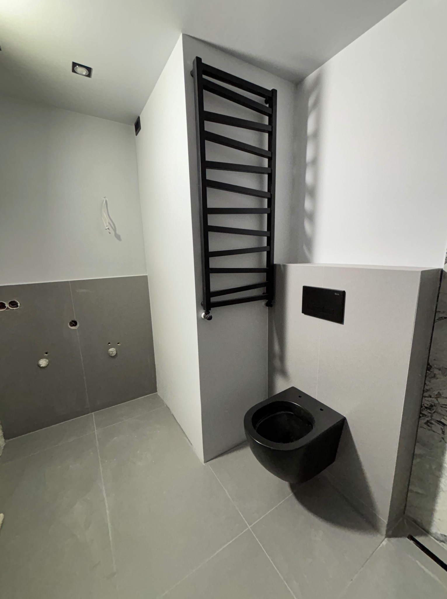 Częściowo wykończona łazienka z czarną wiszącą toaletą, grzejnikiem drabinkowym i szarymi płytkami, widoczne niedokończone instalacje wodne.