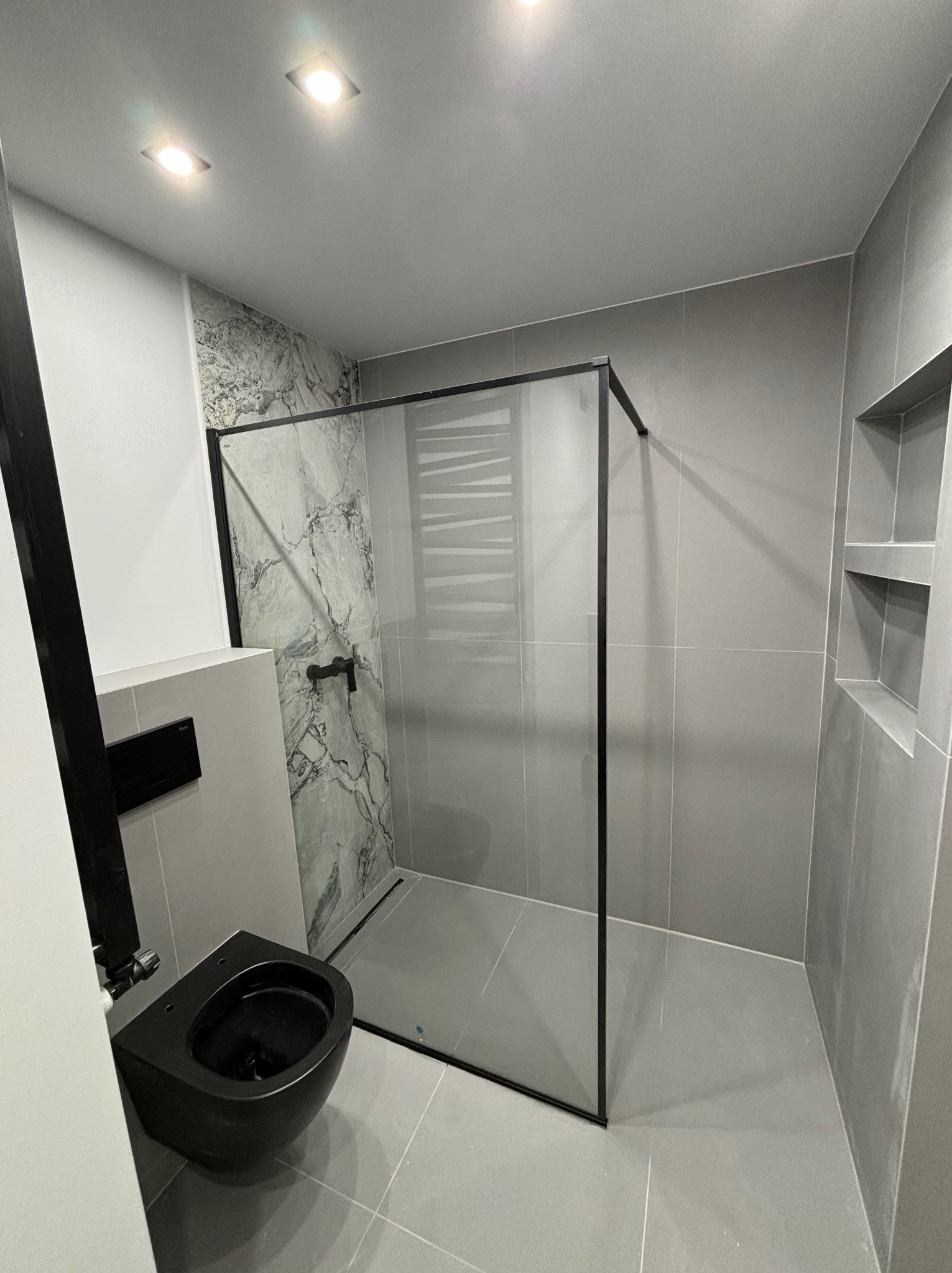 Nowoczesna łazienka w odcieniach szarości z czarną armaturą, kabiną prysznicową typu walk-in z marmurową ścianą i podtynkową toaletą.