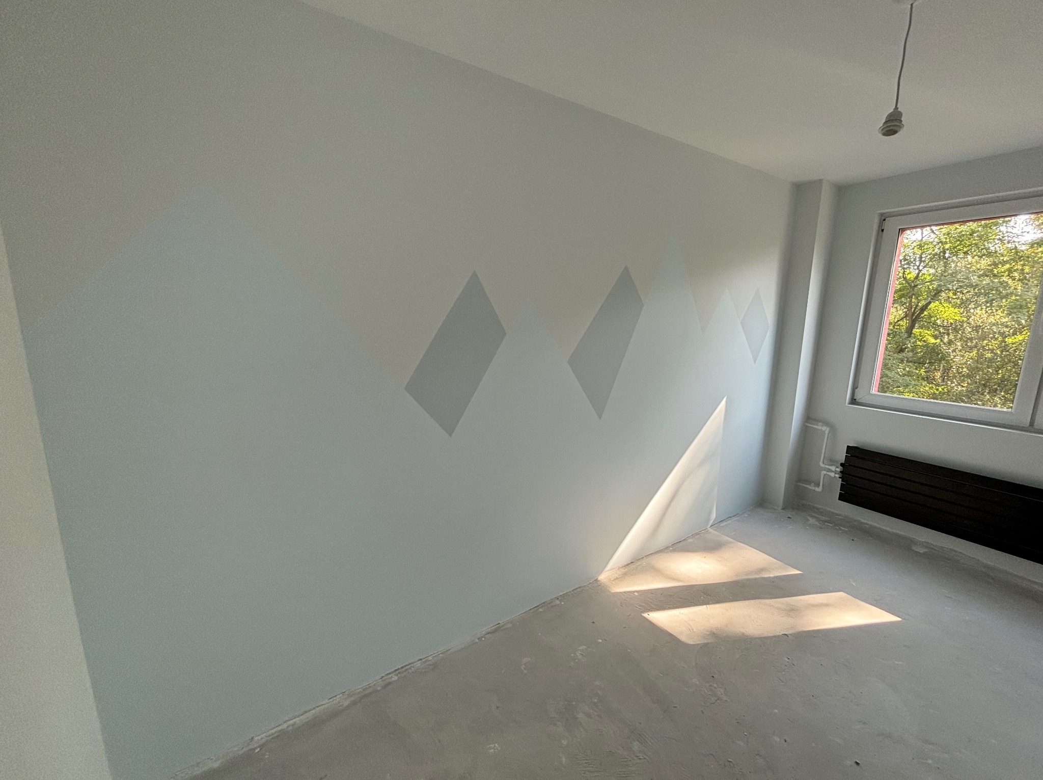 Pokój po malowaniu z motywem gór na ścianie, widoczne promienie słoneczne wpadające przez okno i surowa podłoga.