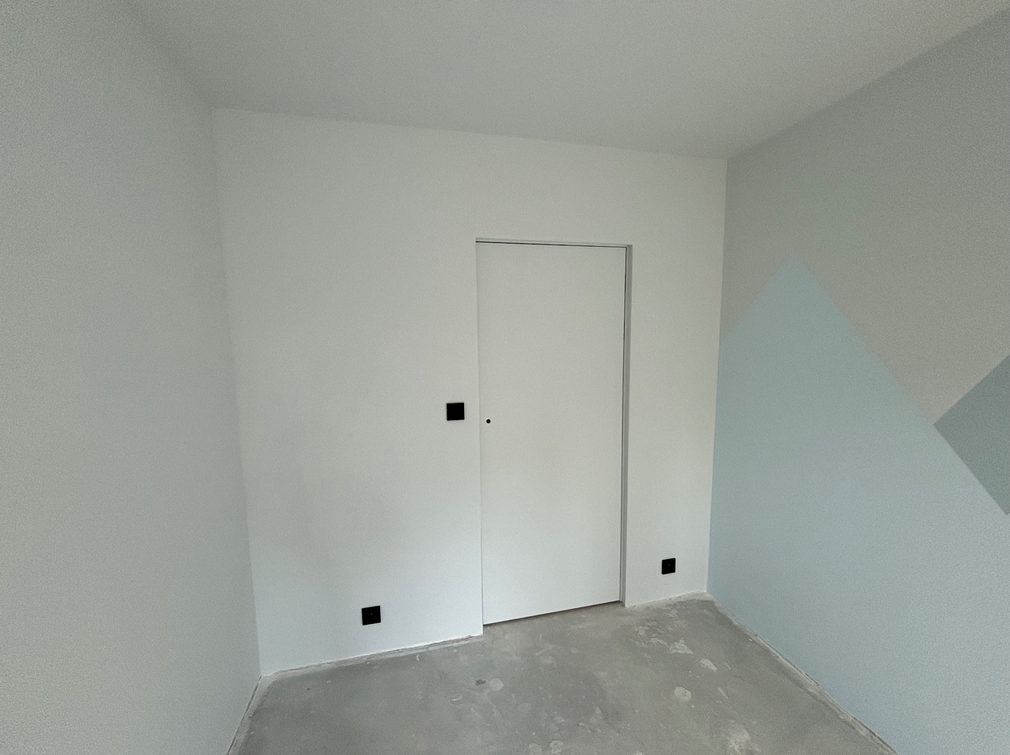 Pomalowane na biało wnętrze pokoju z ukrytymi drzwiami i szarą podłogą, fragment ściany w odcieniach błękitu z geometrycznym wzorem w rogu.