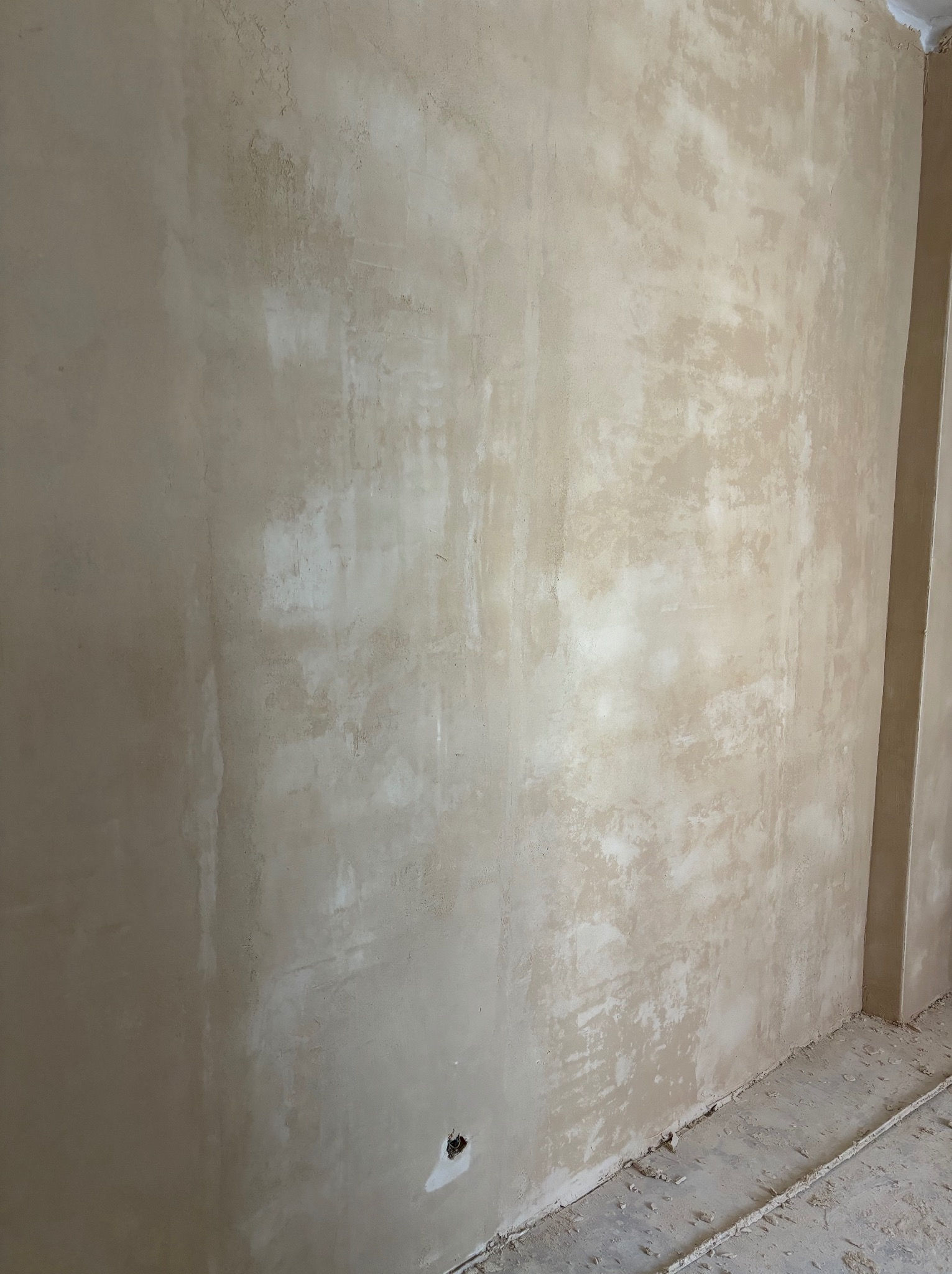 Ściana po nałożeniu pierwszej warstwy tynku w odcieniach beżu, widoczne nierówności i surowy stan wykończenia, z otworem na instalację elektryczną w dolnej części.