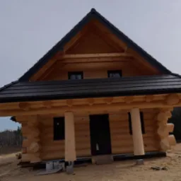 Surowy stan drewnianego domu z bali z czarnym dachem z blachy trapezowej, widoczne niezabezpieczone podpory ganku i teren budowy.