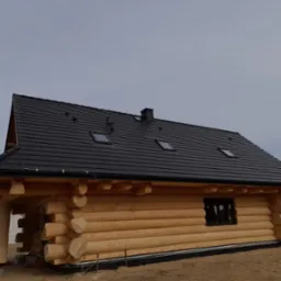 Nowo wybudowany, drewniany dom z bali z czarnym, skośnym dachem pokrytym dachówką modułową i trzema oknami dachowymi, usytuowany na działce z widocznym fragmentem lasu w tle.
