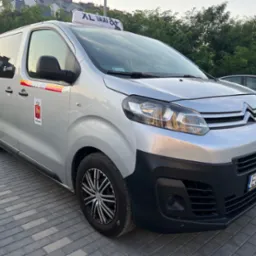 Srebrny van z oznaczeniami XL Taxi 8+ na dachu, zaparkowany na brukowanej nawierzchni, z widocznym herbem Warszawy i numerem telefonu na boku.