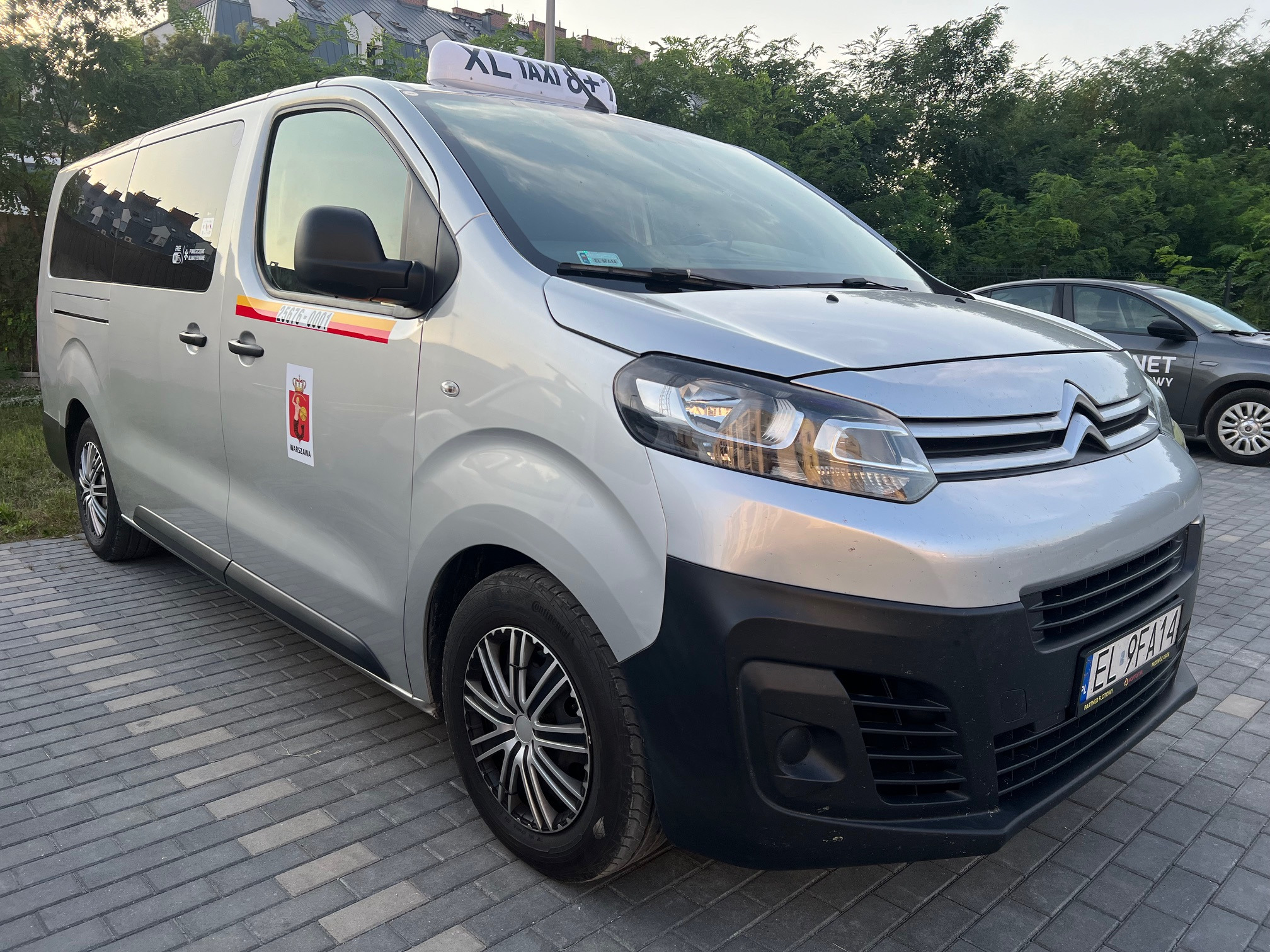 Srebrny van z oznaczeniami XL Taxi 8+ na dachu, zaparkowany na brukowanej nawierzchni, z widocznym herbem Warszawy i numerem telefonu na boku.