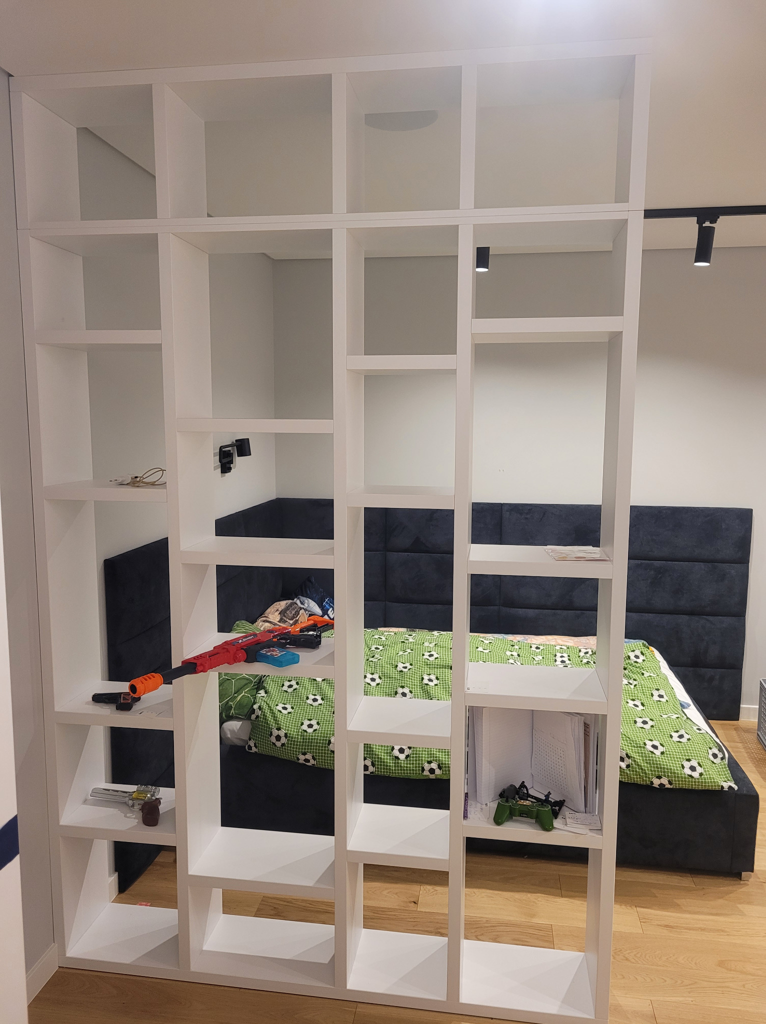 Biały regał na książki, na tle łóżka z zieloną pościelą w piłki. Regał z licznymi półkami, częściowo wypełniony przedmiotami.