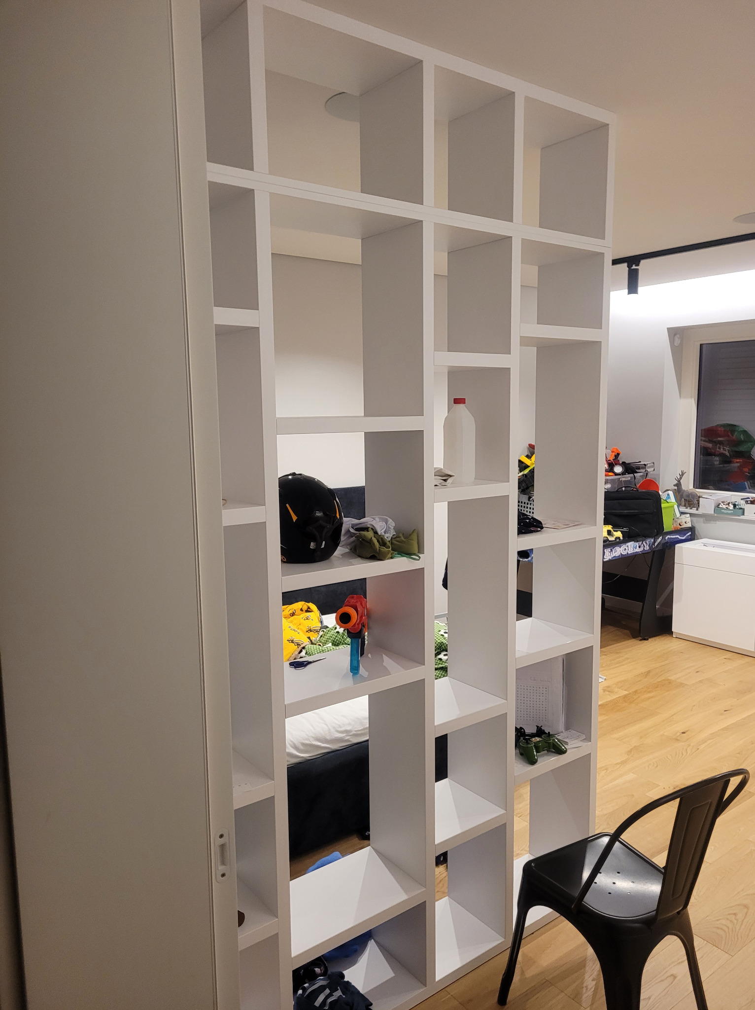 Biały regał z kwadratowymi półkami, częściowo zasłaniający widok na pokój dziecięcy z łóżkiem i biurkiem. Obok stoi czarne krzesło.