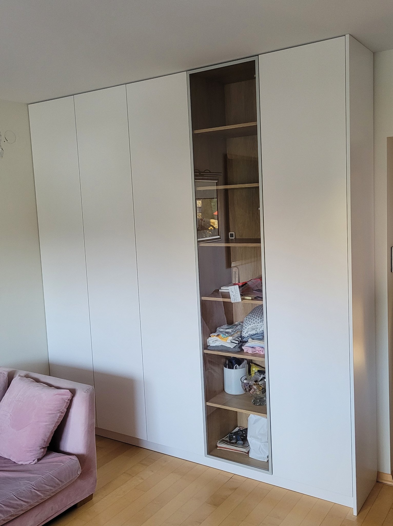 Biała szafa wnękowa z otwartą półką, widoczne ubrania i przedmioty na półkach. Minimalistyczny design, drewniana podłoga, fragment różowej sofy.