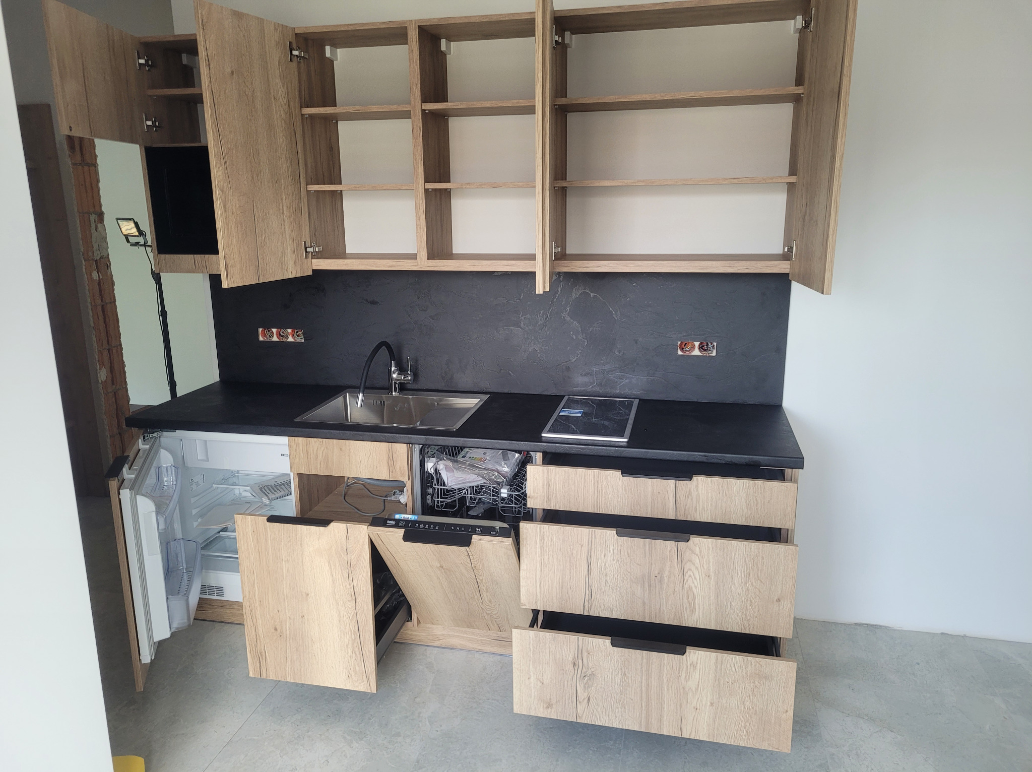 Jasna kuchnia z drewnianymi szafkami, czarnym blatem i otwartymi drzwiczkami lodówki i zmywarki, ukazująca detale wyposażenia.