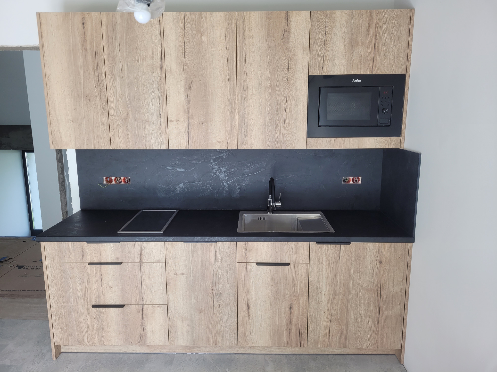 Jasna zabudowa kuchenna z drewnianymi frontami i czarnym blatem, z wbudowaną mikrofalówką Amica. Realizacja w Nienadówce.