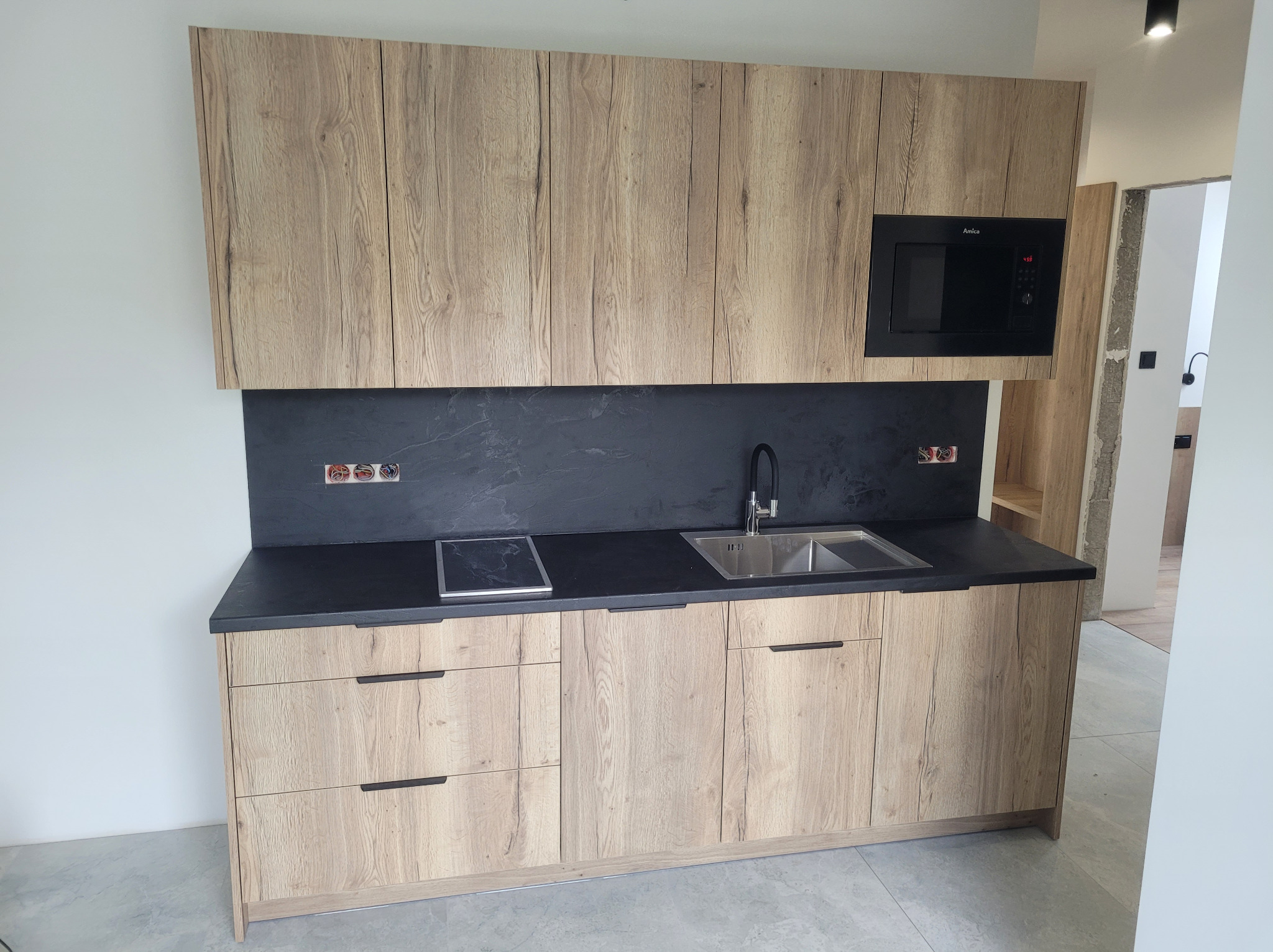 Jasna kuchnia z drewnopodobnymi frontami, czarnym blatem i płytą indukcyjną. W tle widoczny fragment innego pomieszczenia.