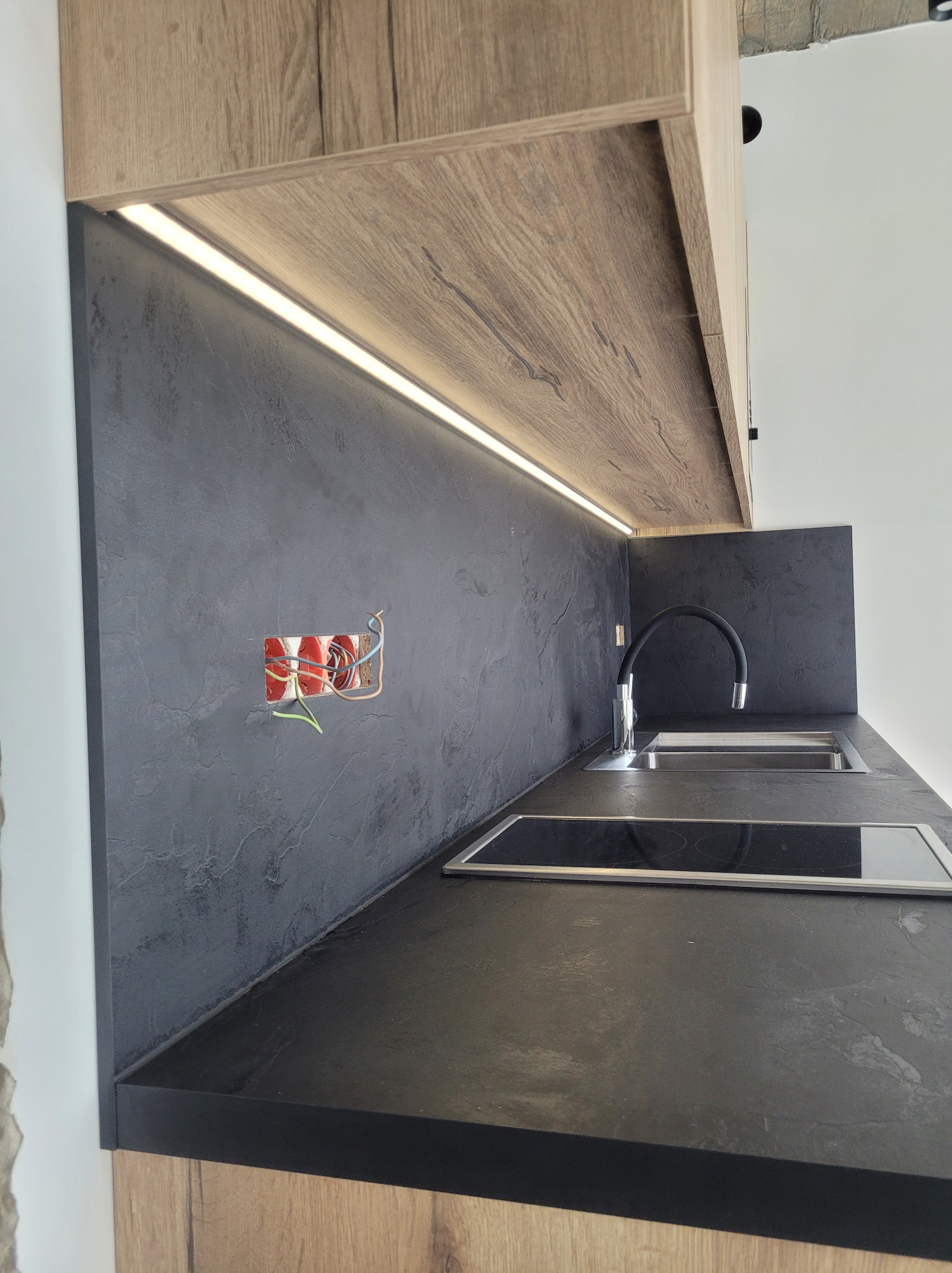 Częściowo wykończona kuchnia: czarny blat i ściana, drewniane szafki, oświetlenie LED, zlew i płyta indukcyjna. Widoczne kable elektryczne w ścianie.
